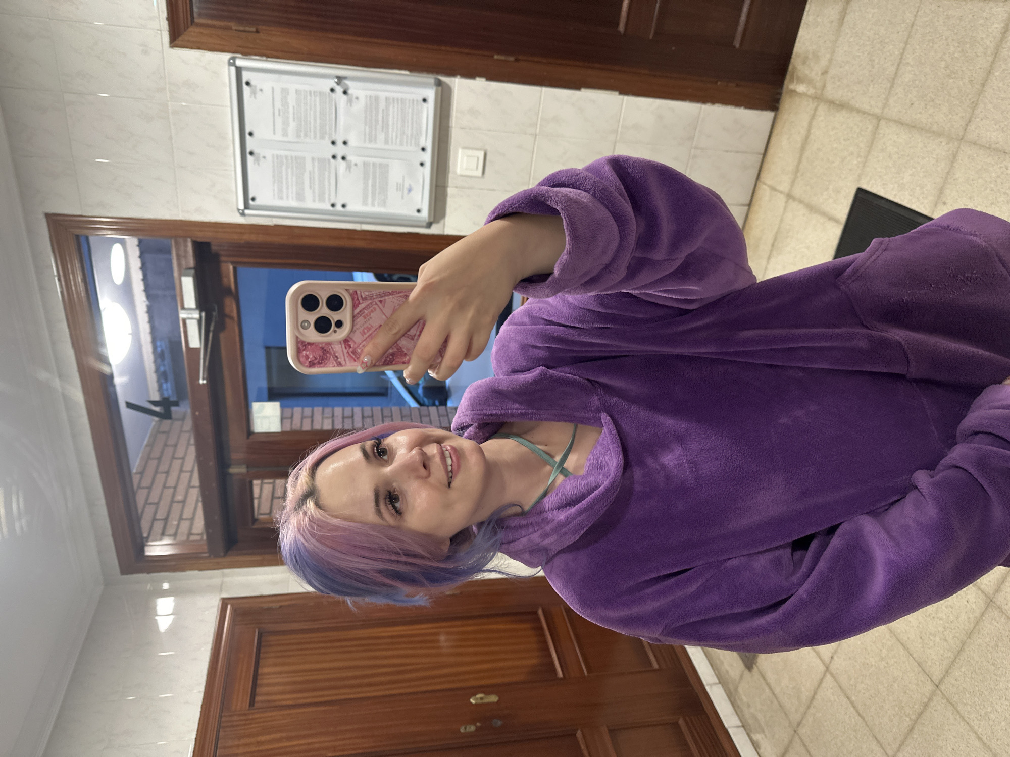 The image shows a young woman taking a selfie, holding a smartphone with a pink case in her right hand. She is smiling and looking towards the camera. Her hair is dyed in shades of purple and blue. She is wearing a fluffy, hooded, purple garment, possibly a robe or a hoodie, over a light blue top.
The setting appears to be an indoor hallway or common area. The background features a white tiled wall with wooden door frames and a window revealing an outdoor paved area with a brick pattern, suggesting it might be evening or night outside. A bulletin board is mounted on the tiled wall, displaying four sheets of paper with illegible text. Below the bulletin board, a light switch is visible. To the right, part of a wooden door with a brass handle is seen. The floor is tiled, featuring light-colored, possibly off-white or beige, tiles, with a dark mat near the bottom right of the frame. The overall impression is a cozy indoor moment.