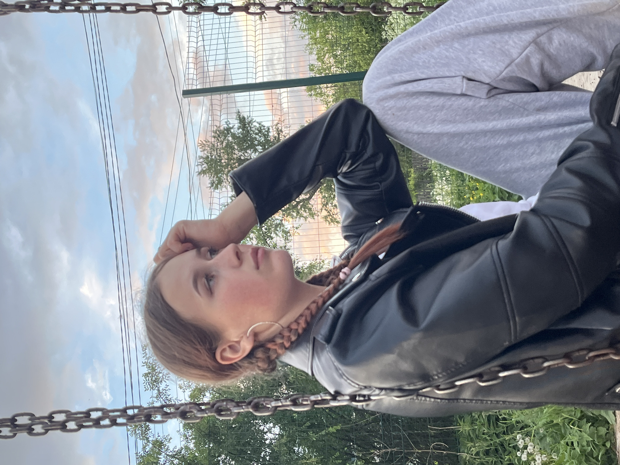 A young woman is sitting on a swing, outdoors. She is wearing a black leather jacket and grey sweatpants. Her brown hair is styled in braids. A chain fence is visible in the background, along with trees and a cloudy sky. The location cannot be determined from the image.