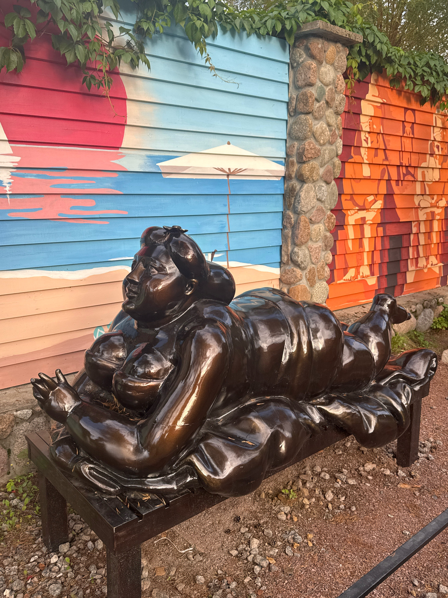 A bronze sculpture of a reclining, voluptuous woman is situated on a dark wooden bench. The sculpture is positioned outdoors in front of a vibrantly painted wooden wall. The wall features a mural on the left depicting a beach scene with a red sun, blue sky and water, and a sandy shore, along with a white beach umbrella. To the right of the beach mural, the wall transitions to a section with a stone pillar, and then to another mural with orange and red tones, featuring stylized human figures. Green vines with leaves cascade from the top of the wall and the stone pillar. The ground around the bench is covered with gravel and small stones. The overall impression is of an artistic installation in an outdoor public space, possibly a garden or a recreational area, located in Интурист Снт, Russia.