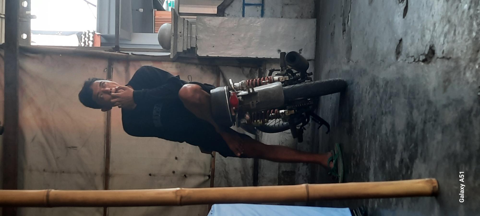 A young Indonesian man, appearing to be in his late teens or early twenties, is captured in a medium shot, leaning against a motorcycle. He is wearing a black long-sleeved shirt and dark shorts, with green flip-flops on his feet. The man is resting his right elbow on his bent left knee, and his right hand is near his mouth, as if in thought or speaking. His gaze is directed upwards and to his right, with his mouth open.
The motorcycle is a dark color, possibly black or dark grey, with visible red shocks on the rear. Its exhaust pipe is prominent. The scene appears to be outdoors, possibly in a garage or covered outdoor area, with a rough, concrete floor. To the left of the frame, a thick bamboo pole is visible, and behind the man, a textured, light-colored wall or tarp is present. In the background, there is a glimpse of a building with architectural details, suggesting an urban or semi-urban setting. The lighting indicates it is daytime, with natural light filtering into the scene. The overall atmosphere is casual and unposed. The image is stamped with "Galaxy A51" in the bottom left corner, indicating it was taken with a Samsung Galaxy A51 smartphone. The location is identified as Selopuro, Indonesia.