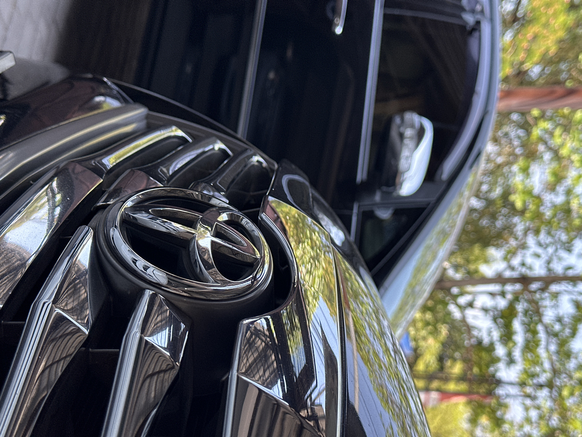 A close-up, low-angle shot focuses on the front grille of a black Toyota car, prominently displaying its chrome emblem. The car is parked outdoors, with a blurred background of green foliage and trees, suggestive of a residential area or park. To the left of the frame, another black car is partially visible, its side mirror reflecting the surrounding environment. The lighting indicates it is daytime. The location is identified as Bulakrejo, Indonesia.