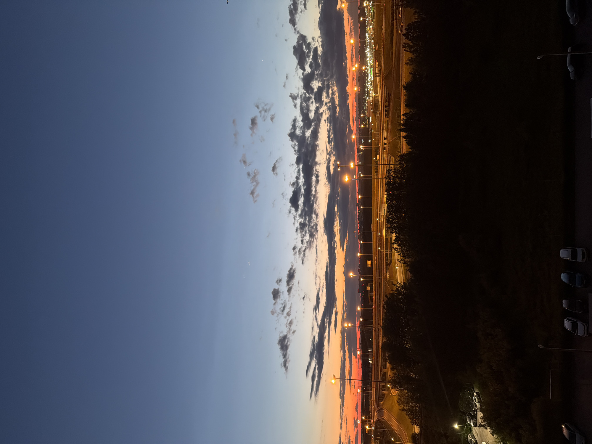 The image captures a twilight scene overlooking a highway interchange in an urban environment. The sky transitions from a deep blue at the top to a vibrant orange and pink hue near the horizon, indicating the sun has recently set. Wispy clouds are scattered across the sky, catching the fading light.
Below the sky, the illuminated highway system dominates the midground. Numerous streetlights line the elevated roads and ramps, casting a warm glow. Vehicles are visible on the highway, appearing as streaks of light due to the long exposure or the twilight conditions.
In the foreground, a dense line of dark trees creates a silhouetted barrier. Beyond the trees, a dimly lit parking area is visible with several cars parked. Further down, at the bottom of the image, another parking area is even darker, with a few cars illuminated by faint light sources. The overall impression is one of urban infrastructure at dusk, with the artificial lights of the city beginning to take over from the natural light of the sky.