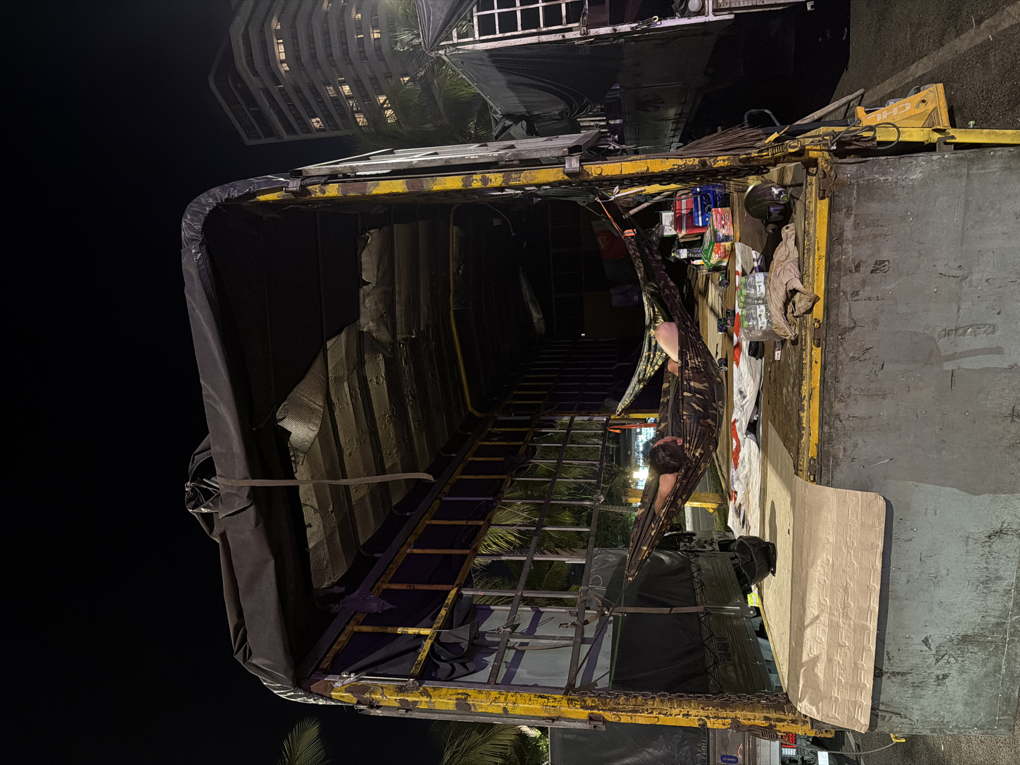 The image depicts the interior of a truck bed parked in Đà Nẵng, Vietnam, at night. The truck bed has a yellow frame and a black tarp partially covering it. Inside, a man is lying in a camouflage-patterned hammock. Various items, including bottles, cans, and clothing, are scattered on the flat surface next to the hammock. A building is visible in the background, with lights suggesting it's occupied.