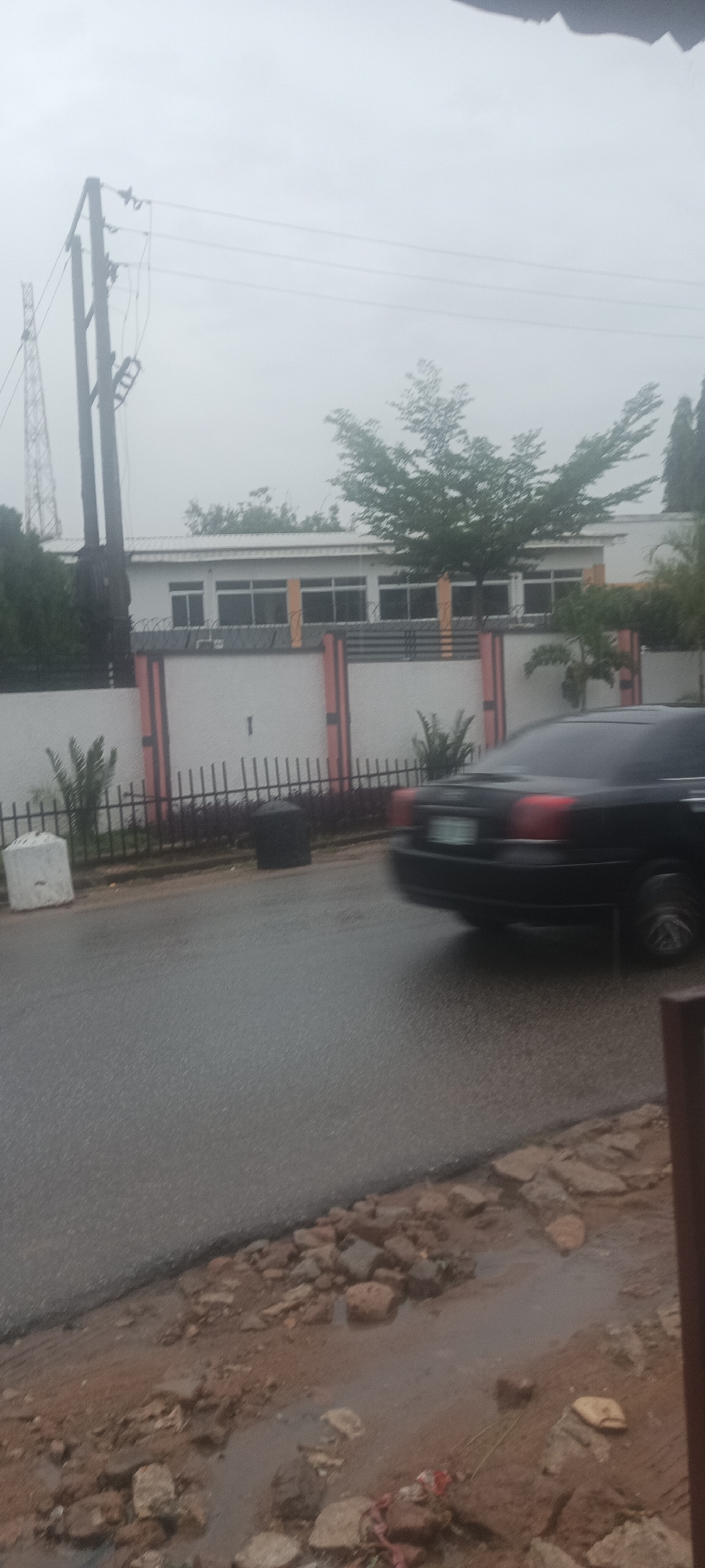 The image shows a street scene in Jos, Nigeria, on a cloudy day. The road is wet, indicating recent rain. A black car is in motion, blurring as it passes by a fence bordering a white building with brown accents. The building has multiple windows. Behind the building, trees are visible. In the background, a power pole and telecommunications tower can be seen. The foreground shows a dirt area with rocks and puddles.