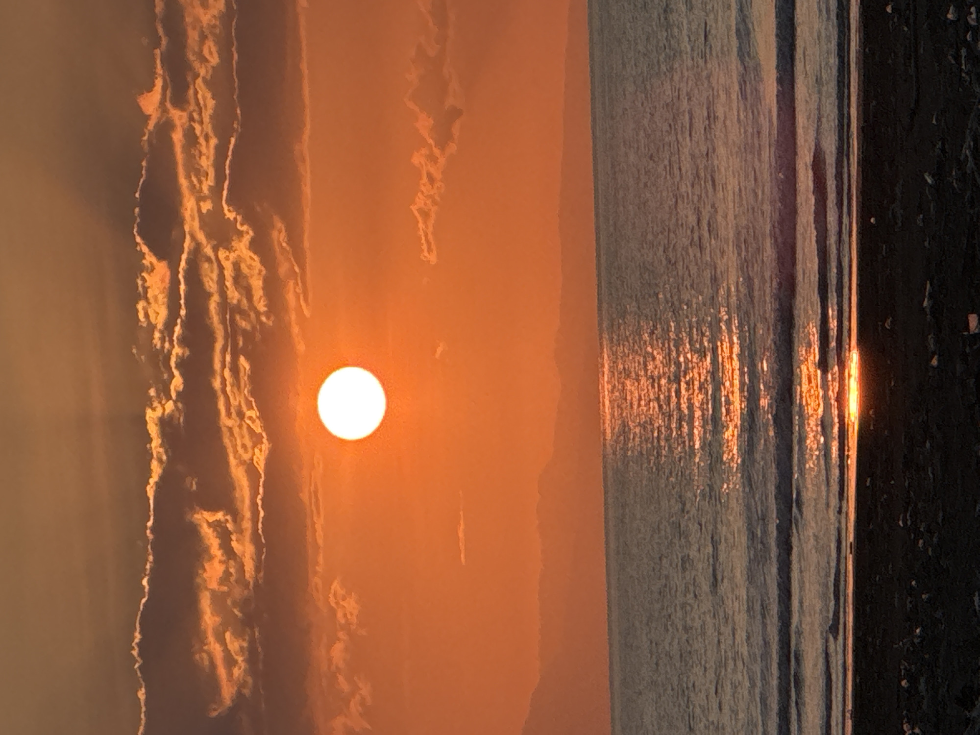 The image depicts a serene sunset over the ocean. The sun, a bright white orb, is positioned high in the sky, casting a warm orange glow across the clouds and water. The clouds are a mix of deep orange and grey, with the sunlight highlighting their edges. In the distance, a faint silhouette of mountains can be seen against the horizon. The ocean is calm, with gentle waves lapping at the shore. The reflection of the sun shimmers on the water's surface, creating a path of light. The foreground shows a dark, sandy beach with scattered debris. There are no people or animals visible in the image, and no discernible structures or man-made objects other than the debris on the beach. The overall impression is one of natural beauty and tranquility. The location is the coastal area of Meunasah Jurong, Indonesia.