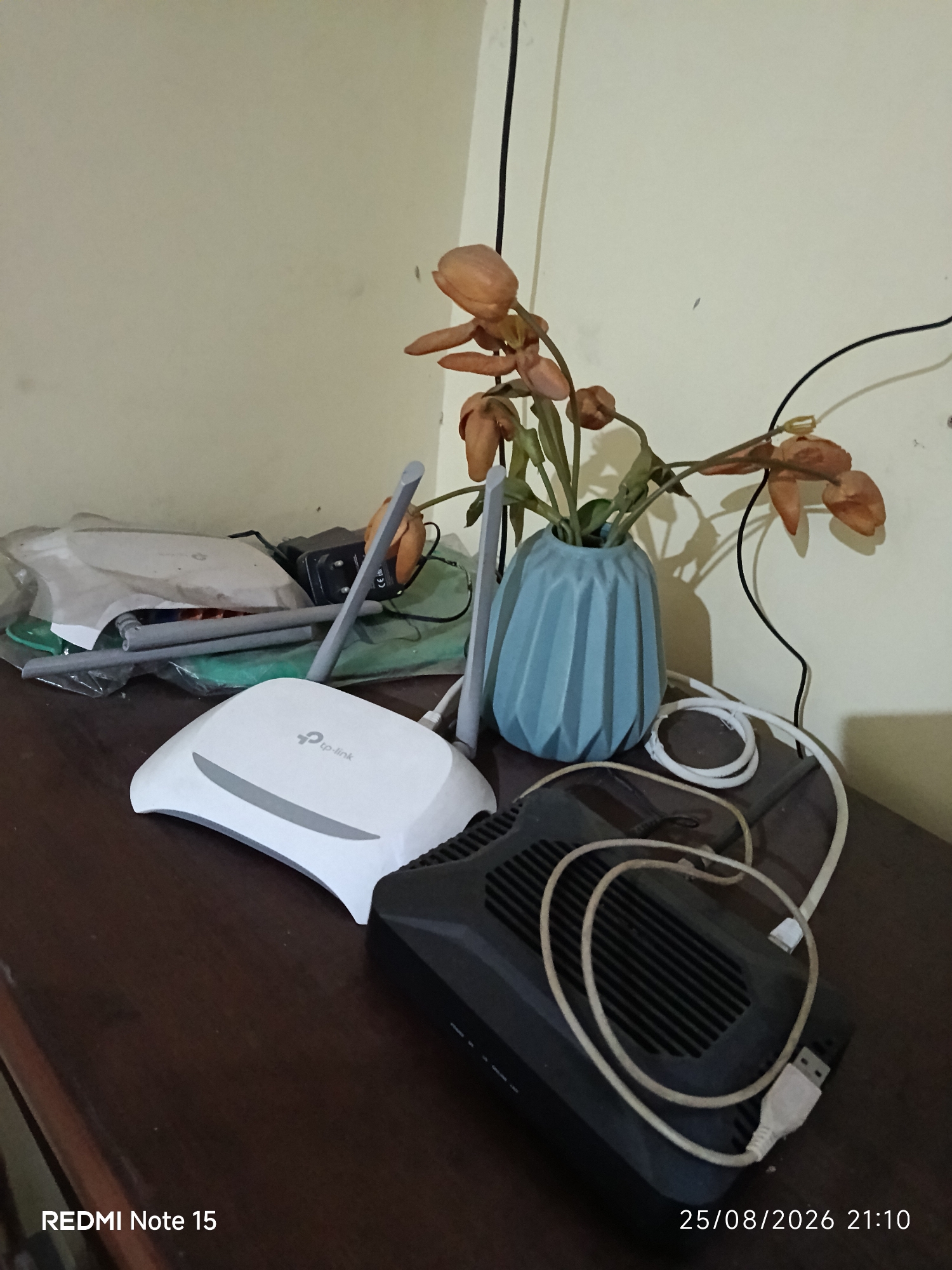 The image shows a wooden table with several objects on it. In the foreground, a white TP-Link router is placed next to a black network device. A white USB cable is coiled and connected to the black device. Behind these, there is a blue vase containing dried flowers. To the left, there are some plastic-wrapped items and a power adapter. The background is a plain wall. The timestamp "25/08/2026 21:10" is visible in the bottom right corner, along with "REDMI Note 15" in the bottom left. The image appears to be taken indoors, likely in a residential setting. There is no information or visual cues in the image to indicate its location in Keboananom, Indonesia.