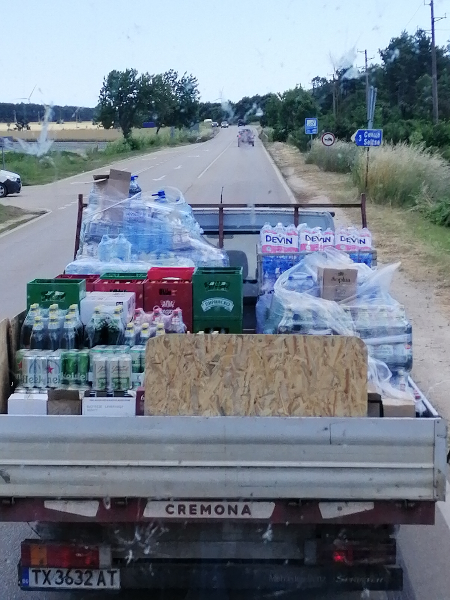 This is a medium shot, taken from a vehicle, of a truck loaded with bottled water and beverages driving down a road. The truck is carrying a significant amount of bottled water, much of it wrapped in plastic, and several crates of beer. Some of the water bottles have "DEVIN" printed on them. There are also some boxes and crates of other items, including what appears to be Heineken beer. The scene is set on a two-lane road with some greenery and trees on the side. In the distance, there are wind turbines visible, suggesting a rural or semi-rural landscape. There are also road signs, one indicating "3 Селце Seltse," which is likely a town name and distance. The sky is overcast, suggesting it might be morning or late afternoon with cloudy weather. The truck itself has "CREMONA" written on the back and a license plate with the characters "TX 3632 AT". The overall impression is of a delivery or transport of goods in what appears to be Bulgaria. There are no people visible in the image. The lighting and the presence of what looks like condensation on the windshield suggest it is daytime, but possibly not bright sunlight.