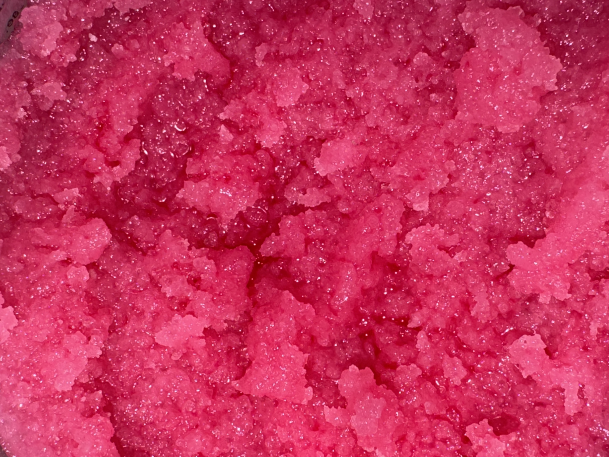 The image is a close-up, overhead view of a vibrant pink, granular substance, likely a sugar scrub or bath bomb mixture. The texture is coarse and uneven, with many small, crystalline particles clumped together. There are also small, glistening highlights suggesting moisture or oil mixed within the granules. The lighting is bright, creating strong reflections and emphasizing the color. The scene is static, with no visible subjects, actions, or interactions. The context of "Яромінскі сельскі Савет, Belarus" is a geographical location and does not pertain to the visual content of the image itself.