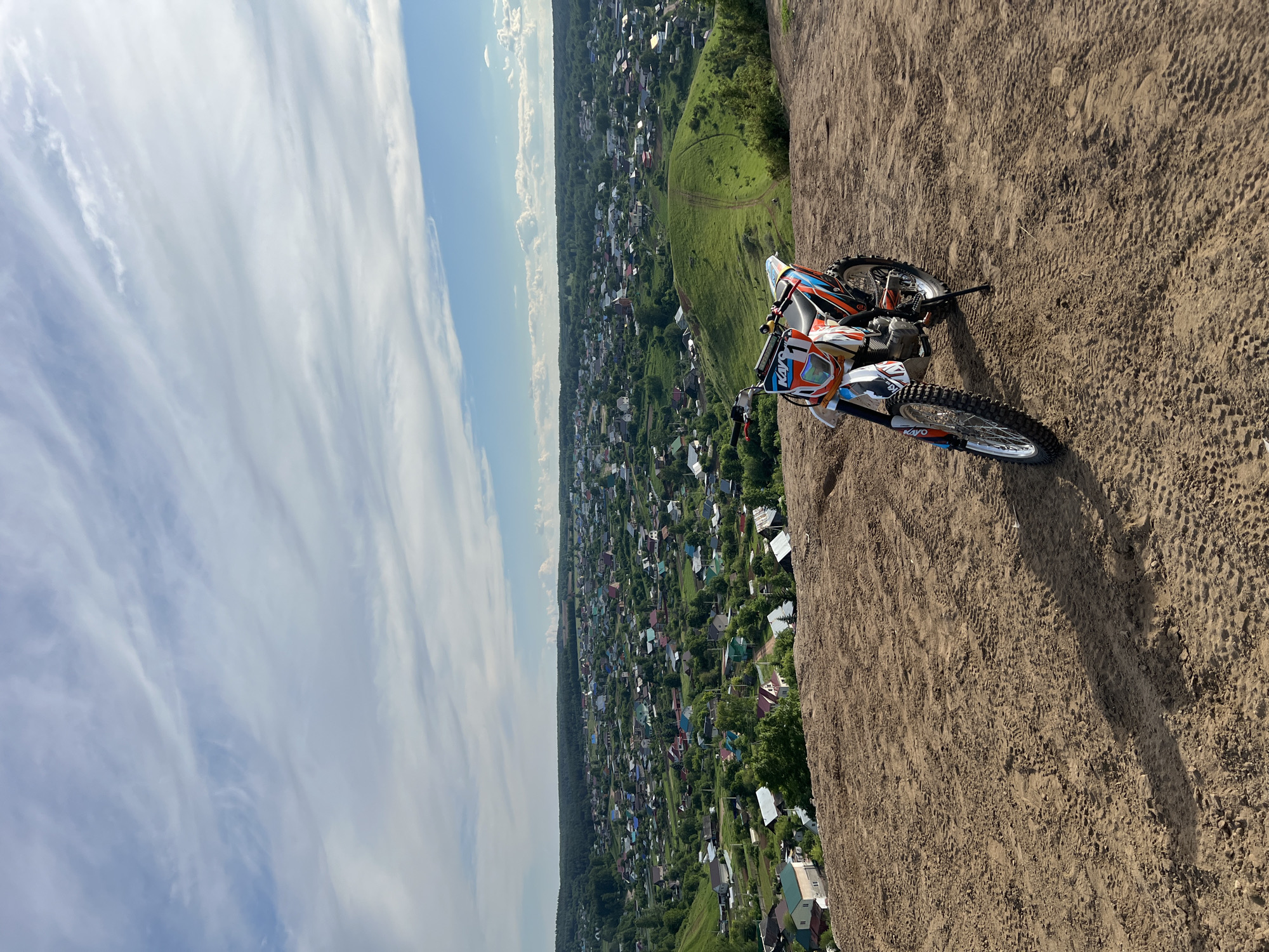 A dirt bike is prominently featured in this image, parked on a sandy, elevated terrain that overlooks a vast valley. The motocross bike is primarily orange, white, and blue, with the brand name "KAYO" clearly visible on its front fairing. It is equipped with knobby tires, indicating its suitability for off-road conditions.
The scene depicts a picturesque landscape in Olykhovka, Russia. Below the elevated position, a densely populated village or town sprawls across the valley, characterized by numerous houses with diverse roof colors, interspersed with lush green trees and vegetation. A faint, winding dirt path can be seen on a verdant hill in the mid-ground. The distant horizon reveals a line of dense forest, suggesting expansive natural surroundings.
The sky is a bright blue, adorned with scattered, wispy white clouds, indicating clear and sunny weather. The lighting suggests it is daytime, likely late morning or early afternoon. No individuals are visible in the image, focusing solely on the bike and the expansive natural and urban landscape. The overall atmosphere conveys a sense of adventure, freedom, and a stunning panoramic view from a high vantage point.