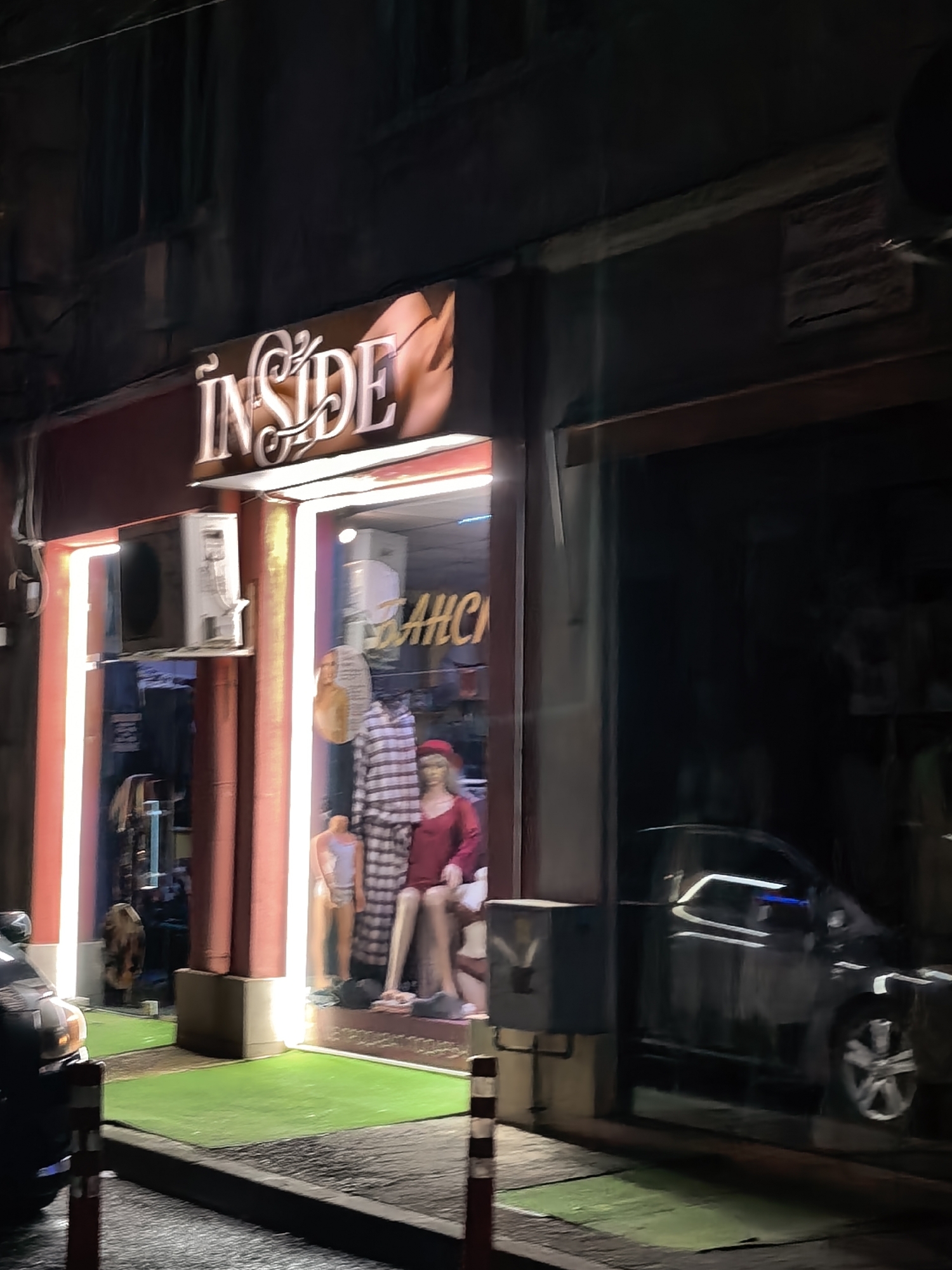 An evening street scene captures a brightly lit shopfront nestled between darker buildings. The main subject is a store named "INSIDE," prominently displayed in stylized, illuminated letters above its entrance. Vertical white light strips frame the shop's glass window, contrasting with the dark street and adjacent structures.
Inside the shop window, several mannequins are visible, showcasing clothing. One mannequin on the left wears a light-colored, possibly striped, two-piece outfit that resembles pajamas or loungewear. Another, more centrally located, is dressed in a vibrant red dress and a matching red beret-style hat. A third mannequin, positioned lower and partially obscured, wears a light blue or lavender top. A small patch of artificial green grass covers the sidewalk directly in front of the shop, extending out to the curb.
To the left of the shop entrance, an outdoor air conditioning unit is mounted on the wall. To the right, the reflection of a dark-colored car, possibly an SUV or hatchback, is visible in the window or a dark recessed entrance. The car's headlights and reflections of streetlights are visible. Two red and white striped bollards stand on the sidewalk in the foreground, near a utility box on the right. The overall atmosphere suggests an urban setting at night, with the shop serving as a beacon of light amidst the darkness. Inside the shop window, part of a word, "BAHÇE," can be discerned.