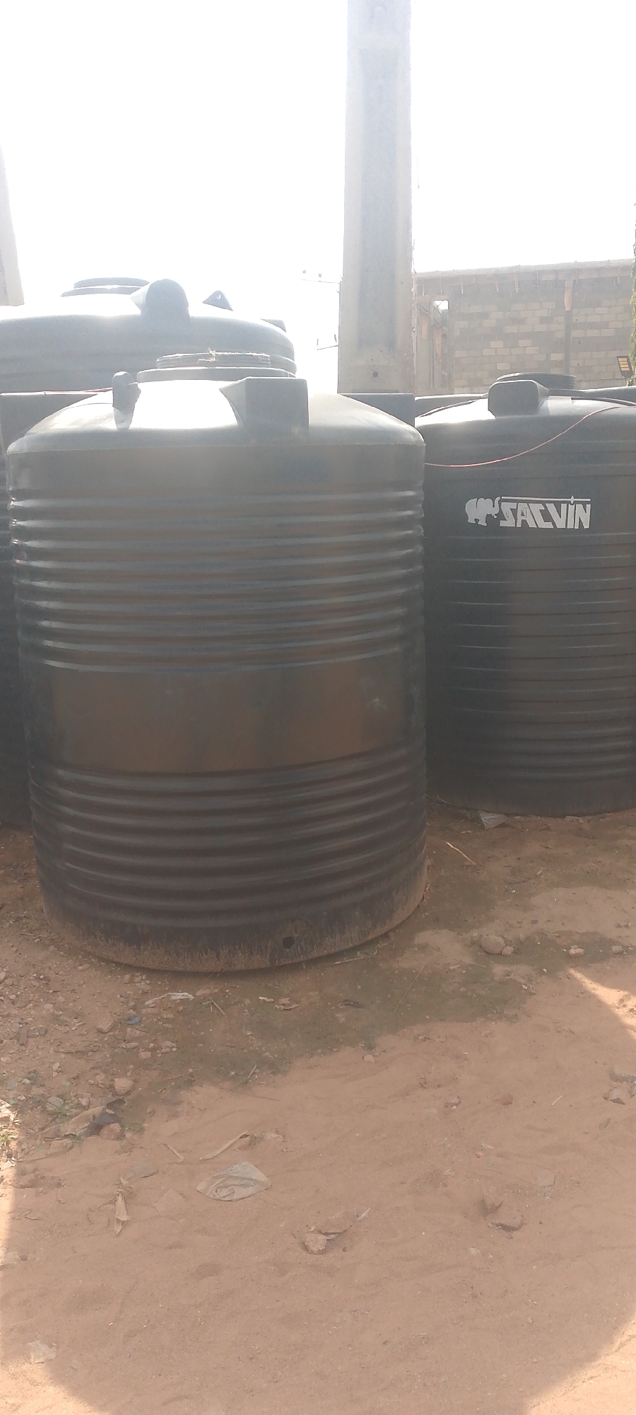 In Jalingo, Nigeria, a sunlit outdoor scene features several large, black, ribbed plastic water tanks arranged on sandy ground. The tanks are cylindrical and have lids with access points. One tank in the foreground is dominant, with its ribbed texture clearly visible. To its right, another tank is partially visible, bearing a white logo with an elephant and the text "SALVIN". The background reveals a partially constructed brick building and a tall concrete pillar, suggesting an industrial or storage area. The ground is covered in loose sand and scattered debris, indicating an unpaved outdoor environment. The lighting is bright, suggesting daytime.