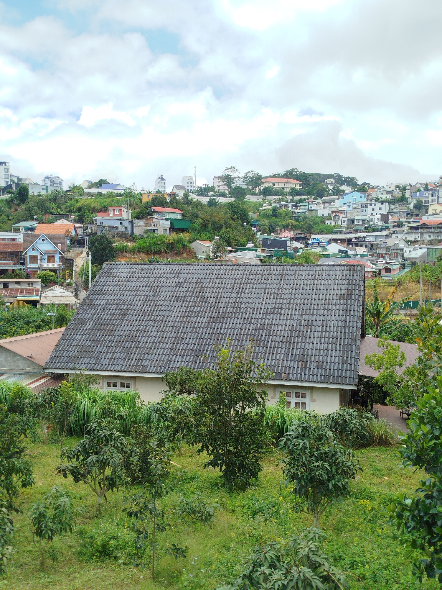 In Da Lat, Vietnam, this image captures a hillside residential area under a cloudy sky. The foreground features a large house with a grey, tiled roof, positioned in front of lush green foliage and smaller trees. Beyond this house, the landscape unfolds into a dense collection of diverse buildings, primarily houses and smaller apartment blocks, built on varying elevations of the sloping terrain. The architecture is varied, with many structures featuring colorful roofs in red, blue, and green, and exteriors in white, cream, and muted tones. The vegetation is abundant, with trees and shrubs interspersed between the buildings, giving the impression of a verdant urban environment. The overall scene depicts a densely populated, hilly residential district on the outskirts of Da Lat.