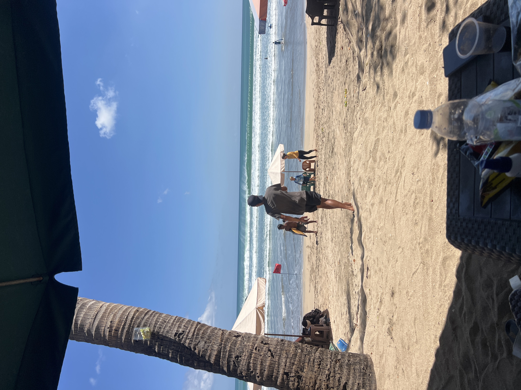 A group of people are enjoying a sunny day at Kuta Beach in Bali, Indonesia. In the foreground, a person wearing a dark t-shirt and shorts walks away from the camera towards the ocean. To the left, a palm tree stands tall, casting a shadow over the sandy beach. Beach umbrellas and chairs are scattered along the shore, providing shade for beachgoers. A few individuals, including two children with a surfboard, are walking near the water's edge. The ocean has moderate waves, and several surfers are visible in the distance. The sky is clear and blue with a few scattered clouds. On a low table in the immediate foreground, a plastic water bottle and a phone are placed.