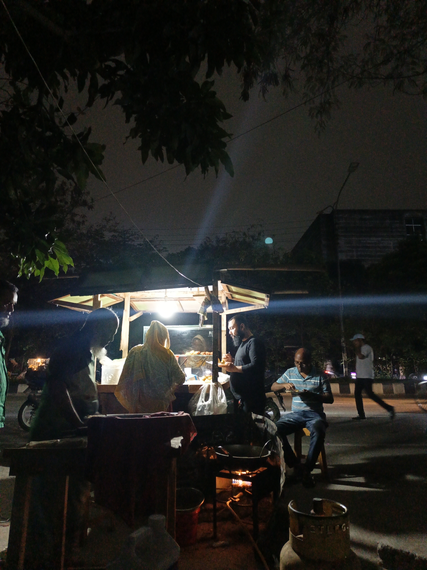 The image depicts a bustling street food stall scene at night in Dhaka, Bangladesh. A brightly lit food cart, likely wooden, forms the central point, illuminated by an overhead light that casts a warm glow on the immediate area while the surroundings remain dark.
Several individuals are gathered around the stall. A vendor, possibly a woman, clad in light-colored or yellow attire with a head covering, stands behind the counter, attending to customers. Food items are visible behind a glass display on the counter, with a faint "K" discernible on the glass. To the right of the vendor, a bearded man in a dark shirt is standing and eating from a plate. Another man, wearing a striped blue and white shirt and jeans, is seated on a small wooden stool, also consuming food.
In the foreground, a cooking station is visible, with a large wok or pan resting over an open flame, indicating fresh preparation. A metallic gas cylinder, bearing some indistinct text, sits near the cooking area. On the left, a man in a green shirt is partially visible, while in the mid-ground on the right, a person in a white cap walks past, and a motorbike is parked.
The scene is set against a dark night sky, with silhouetted tree branches hanging overhead. In the distance, a street light glows faintly, and a dark building stands in the background. The bright illumination of the food stall starkly contrasts with the general darkness, highlighting the activity around it. The overall atmosphere suggests an informal, lively evening meal or snack experience.