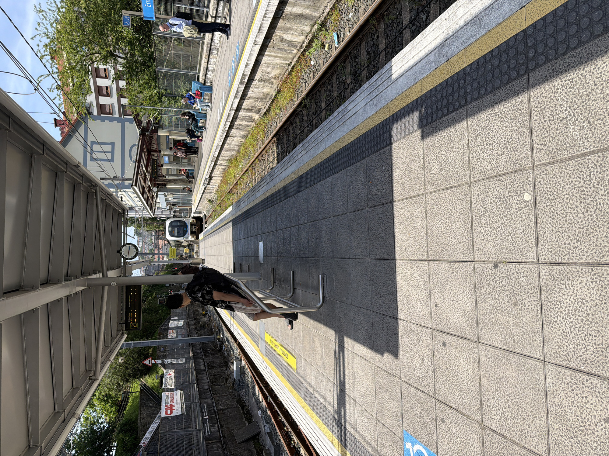 The image shows a train station platform in Errenteria, Spain. The platform is made of gray tiled squares. A person sits on a metal bench. A train is approaching in the distance. Across the tracks, people are sitting on benches on the opposite platform. Buildings and trees are visible behind the opposite platform. There are train tracks running between the two platforms. The image is rotated 90 degrees to the right.