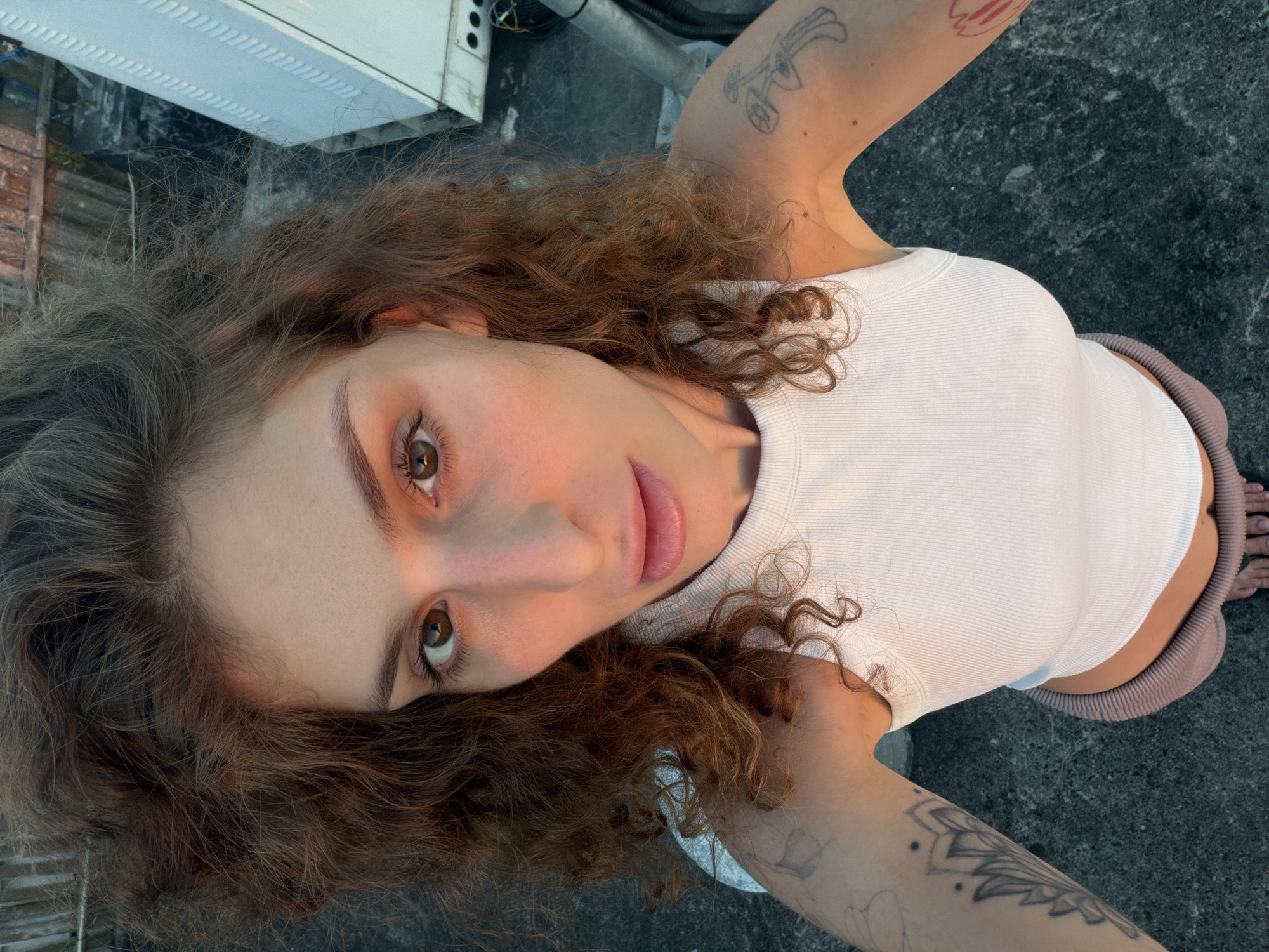 A fair-skinned person with curly brown hair is facing the camera, wearing a white ribbed tank top and mauve-colored pants. The person has visible tattoos on both arms. The location appears to be an outdoor urban setting, potentially in North Kuta, Indonesia, with a gray concrete surface visible and part of a white utility structure in the upper left corner.