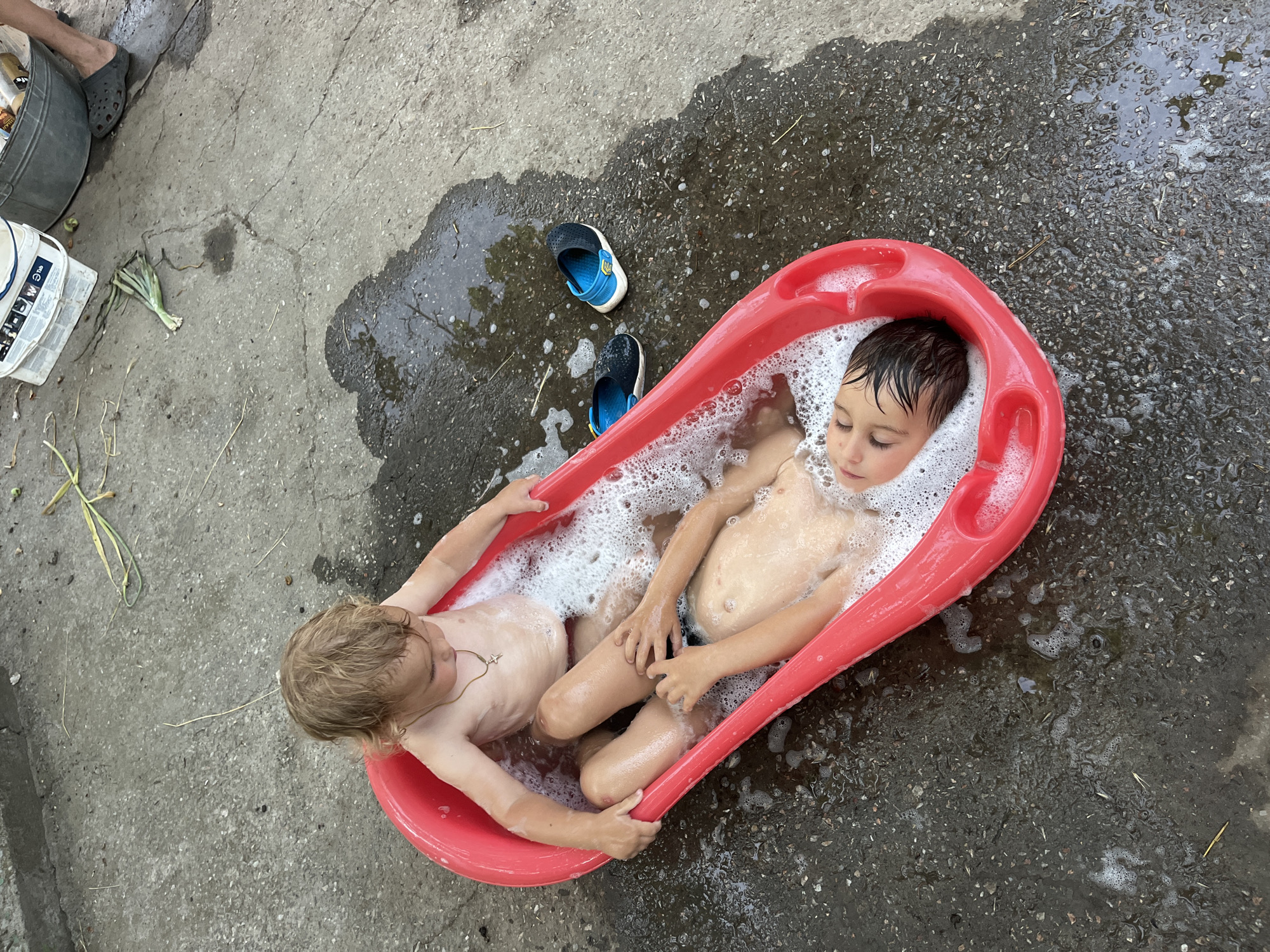Two young boys are bathing in a red plastic tub filled with soapy water on a concrete surface. The tub is positioned outdoors, with scattered debris and puddles of water on the ground. A pair of blue and black water shoes are placed next to the tub. The scene appears to be a casual, perhaps backyard, setting in Chernove, Ukraine.