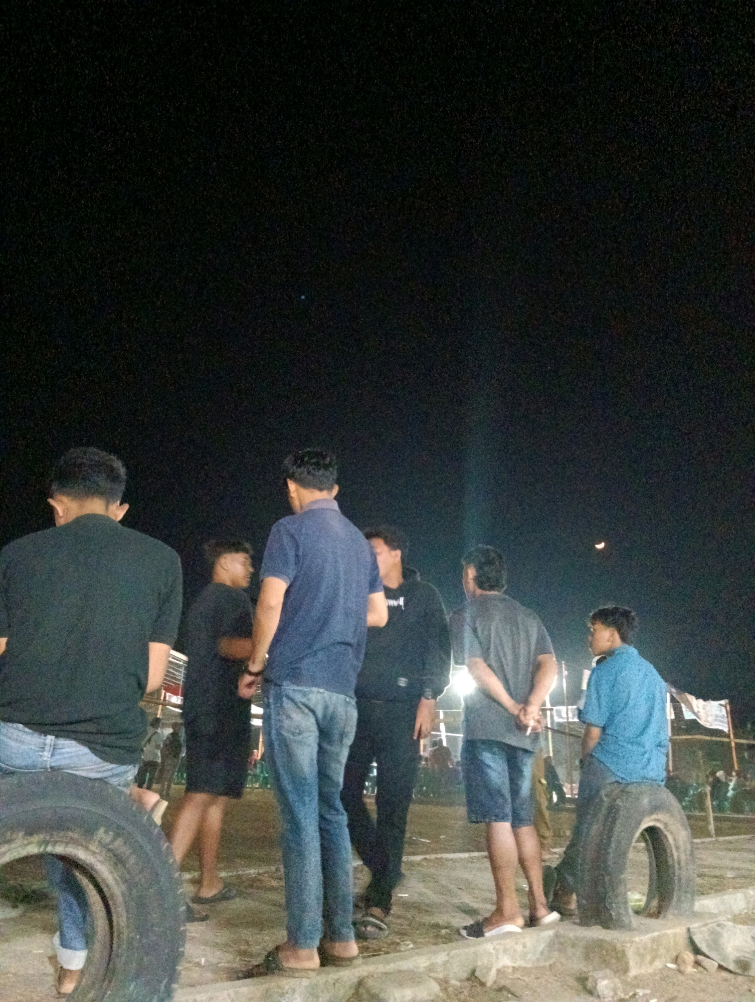 In Pematang Jaya, Indonesia, at night, a group of six young men are gathered in an outdoor area. The scene is dimly lit, with the primary light source appearing to be artificial. The ground is uneven, composed of dirt and some concrete structures.
In the foreground, a large, worn tire lies on its side, serving as a makeshift seat for a young man wearing a black t-shirt and blue jeans. To his left, another young man in a black t-shirt is partially visible, also seated on a tire.
Further into the scene, three men are standing and conversing. One man, wearing a blue t-shirt and blue jeans, stands center-left. To his right, a man in a black hoodie and dark pants is also standing, looking away from the camera. Next to him, a man in a grey t-shirt and blue shorts is standing with his hands behind his back, holding a cigarette.
To the far right, another young man in a blue shirt is seated on a tire, facing away from the camera. In the background, faint structures and banners suggest a public gathering or event space, with other people dimly visible in the distance. The night sky above is dark with a sliver of a crescent moon visible.