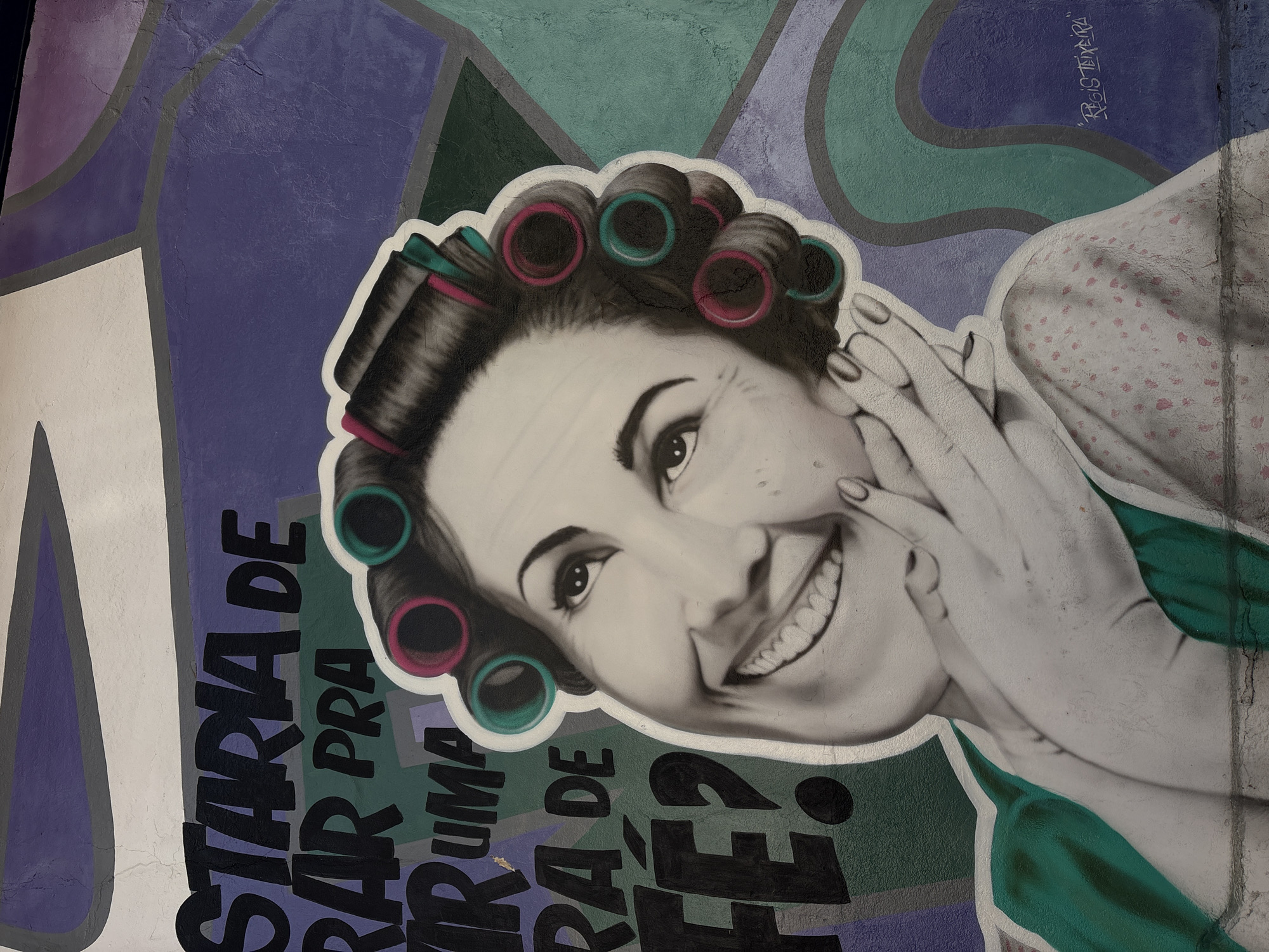 The image is a photograph of a mural painted on an exterior wall in São Paulo, Brazil. The mural features a large, stylized portrait of a smiling woman with hair rollers in her dark hair. The woman is depicted with her hands framing her face, and she is wearing a green top and a patterned dress or shirt. Behind the woman, abstract shapes in purple, green, and white are painted on the wall. Black lettering is visible to the left of the woman's face, appearing to be part of a phrase. A signature, "Regis Teixeira", is visible in the lower right corner of the mural. The wall itself shows some signs of wear and cracking, typical of outdoor urban art.