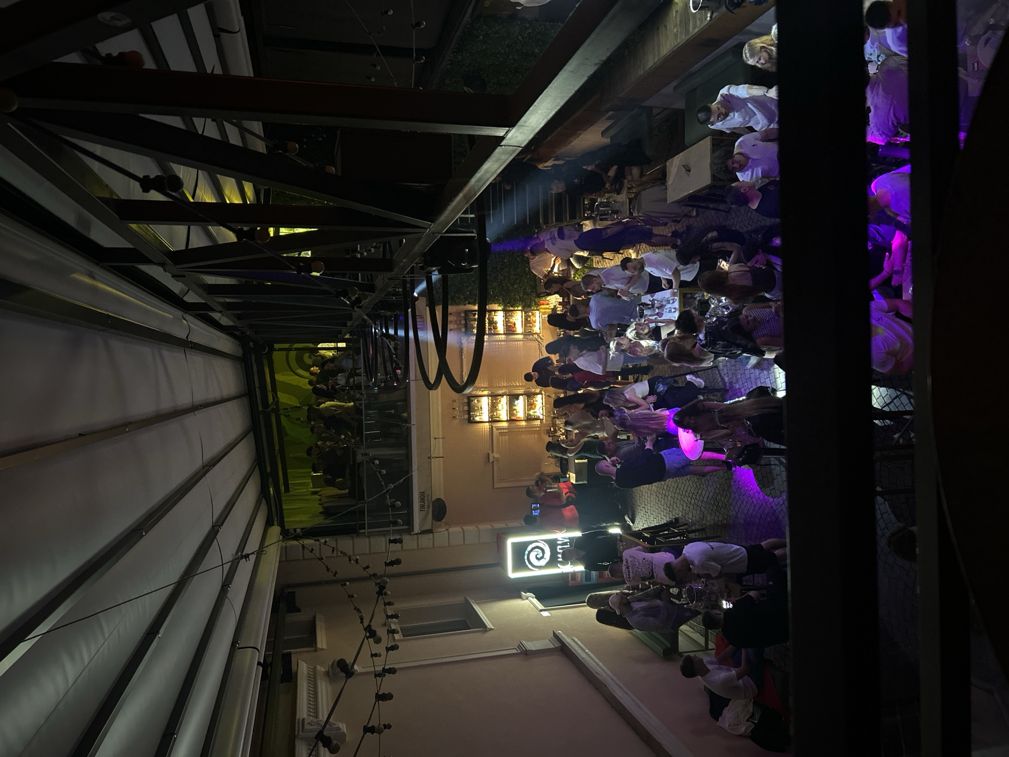 The image depicts an outdoor seating area of a bar or nightclub, likely located in Plovdiv, Bulgaria, given the architectural style and outdoor setting. It is evening, and the space is illuminated by artificial lighting, including string lights, neon signs, and colored spotlights creating a vibrant atmosphere.
In the foreground, several groups of people are gathered around tables, engaged in conversation and socializing. Some are standing, while others are seated. The attire suggests a casual, social gathering. Drinks are visible on some of the tables.
Further into the scene, there is a bar area with patrons interacting with staff. Behind the bar, shelves are stocked with bottles, indicating a well-equipped establishment. Multiple levels are present in the venue, with a balcony or upper-level area visible, also populated with people.
The architecture features exposed beams and a retractable roof structure, suggesting a modern design integrated into a more traditional building. The flooring appears to be cobblestone or textured pavement. The overall impression is of a lively social hub.