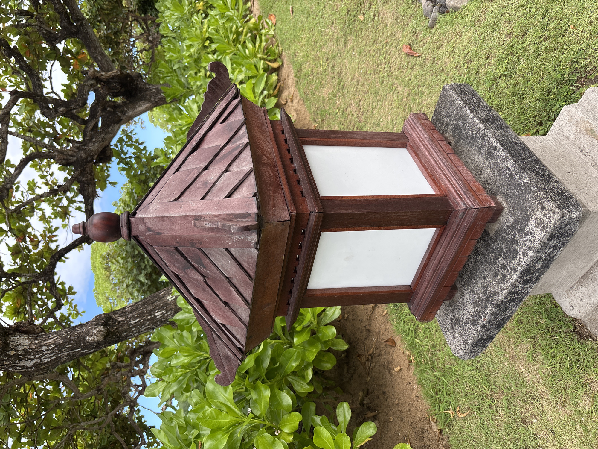 A close-up shot of a wooden Japanese-style lantern on a stone pedestal is set against a backdrop of green foliage and a tree branch, suggesting an outdoor garden setting. The lantern has a multi-tiered, brown-wooden roof structure with layered wooden tiles. The main body of the lantern features white, translucent panels encased in dark wood frames. The lantern sits on a grey stone base. The background includes lush green shrubbery, grass, and the branch of a tree with visible bark texture, and patches of blue sky peaking through the leaves. The location is in Kuta Selatan, Indonesia.