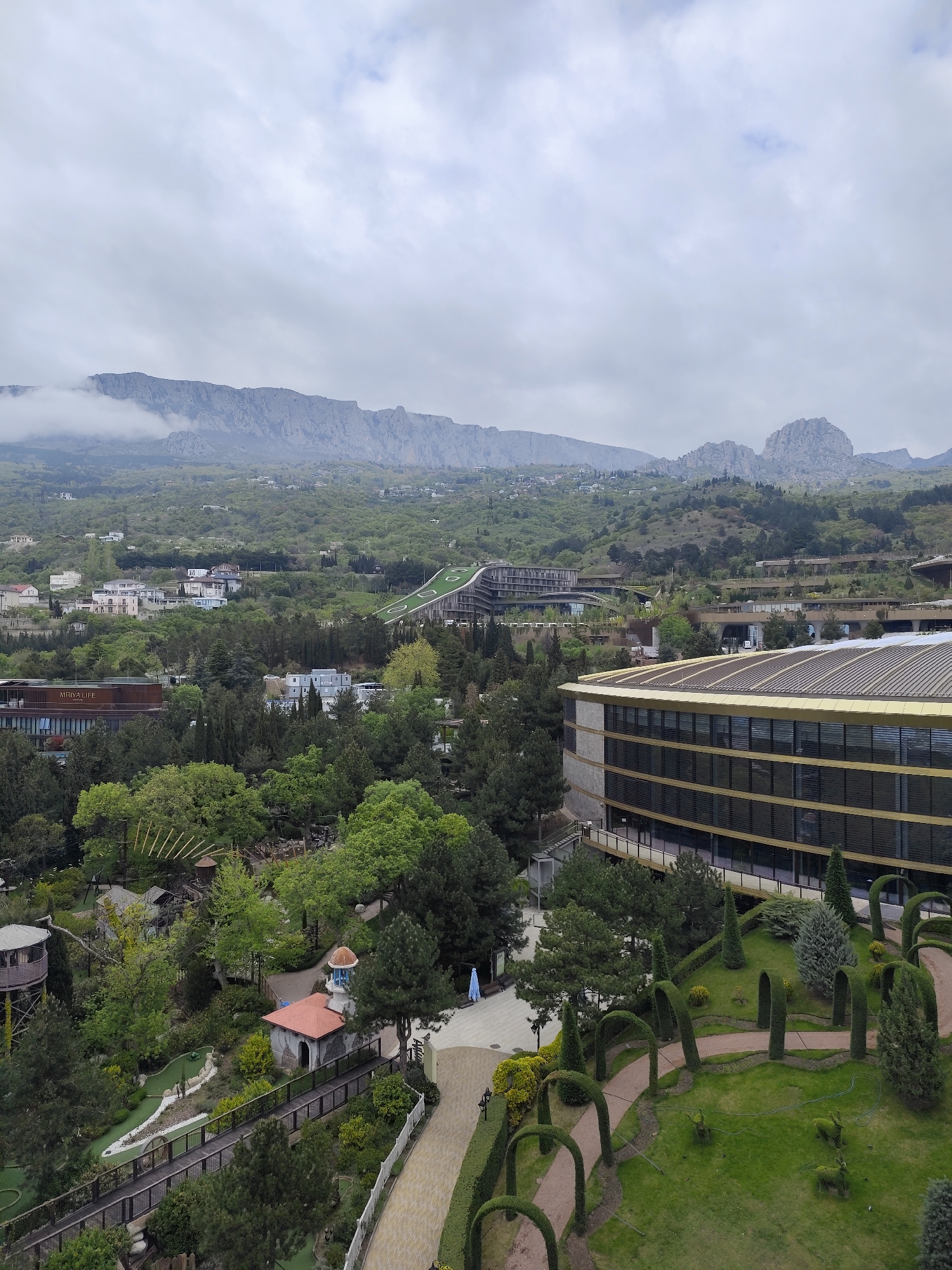 The image depicts a landscape in Симеизский поселковый совет, Ukraine, dominated by a mountain range shrouded in clouds in the background. A mix of architectural styles is seen, including hotels with modern designs and green roofs, interwoven with dense green foliage. A section of a hotel with a gold roof is prominent in the foreground on the right. A miniature golf course and gardens are visible on the left, featuring trimmed hedges and walking paths. The scene is taken from an elevated perspective, likely from a high-rise building. The weather appears overcast. No people are clearly discernible.