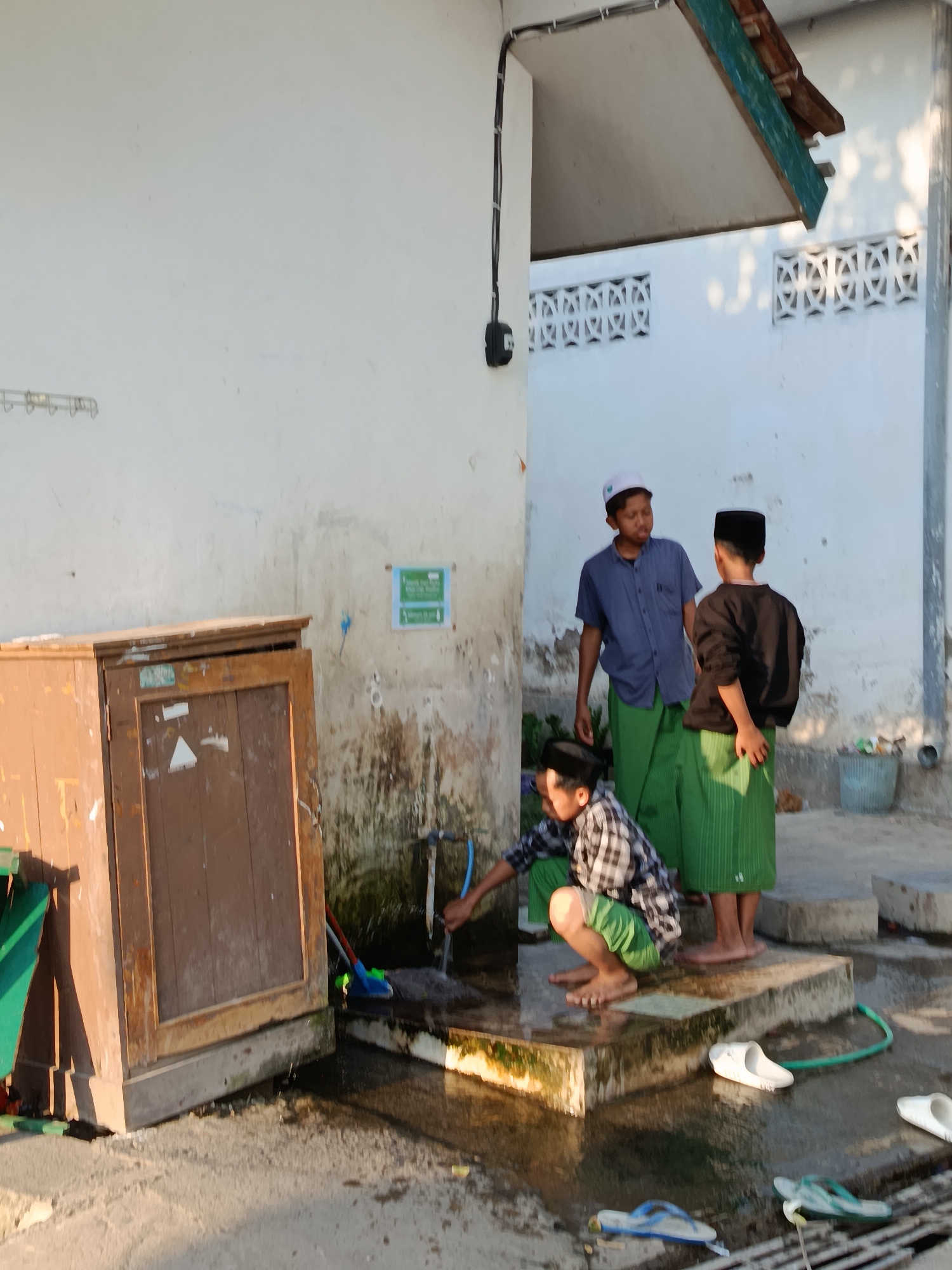 In Ramban Wetan, Indonesia, three young boys are engaged in washing up near a communal water source. The scene is set outdoors, with a white, weathered building providing a backdrop. In the foreground, a wooden cabinet with a weathered door stands to the left. To the right of the cabinet, a concrete structure with a water tap is actively being used. One boy, wearing a checkered shirt and green shorts, is crouched down and washing his hands under the running water. He is barefoot. Two other boys stand nearby, observing. One, in a blue shirt and green sarong, faces the boy at the tap. The other boy, in a black long-sleeved shirt and green sarong, faces away from the viewer, also looking towards the washing area. The ground is wet and reflective, indicating recent water usage. Several pairs of flip-flops are scattered around the wet area. The environment appears to be a shared outdoor space, possibly a mosque or a community area, given the traditional attire of the boys. The lighting suggests it is daytime, possibly late afternoon, with soft shadows present.