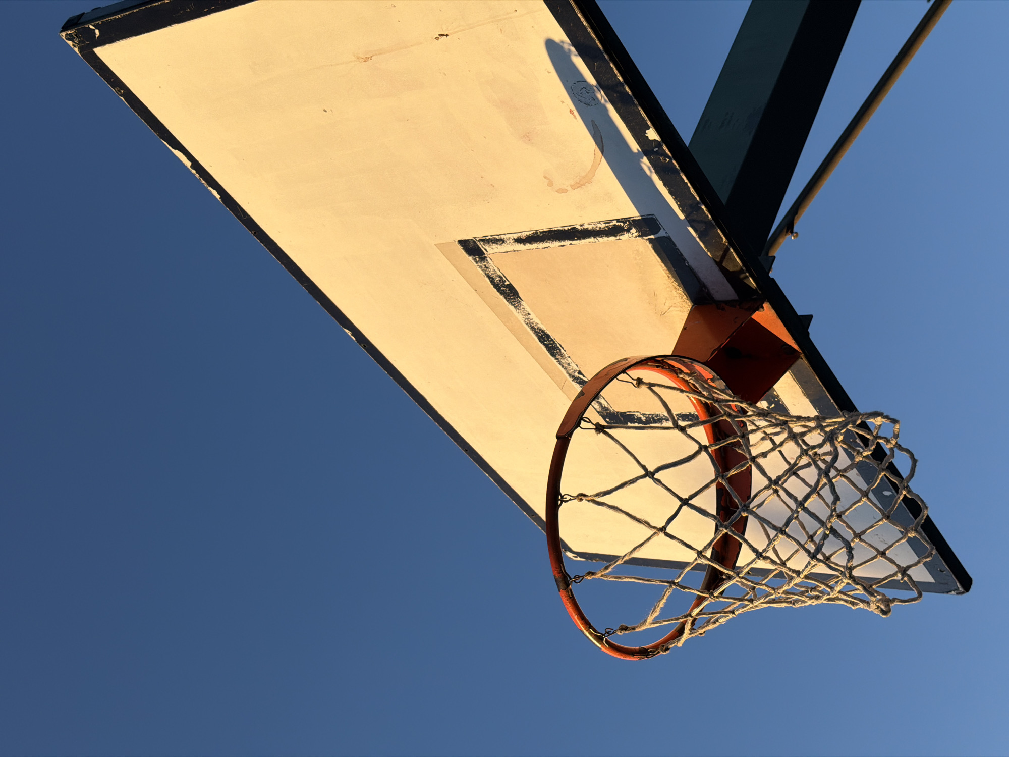 The image shows a basketball hoop and backboard against a clear blue sky. The backboard is white with a black border and a faded square marking, likely for the target area. The hoop is orange and has a white net. The support structure for the hoop and backboard is dark green and metal. The low-angle shot gives the impression that the viewer is looking up at the basketball hoop. The context of the city of Milan is not evident in the photo itself.