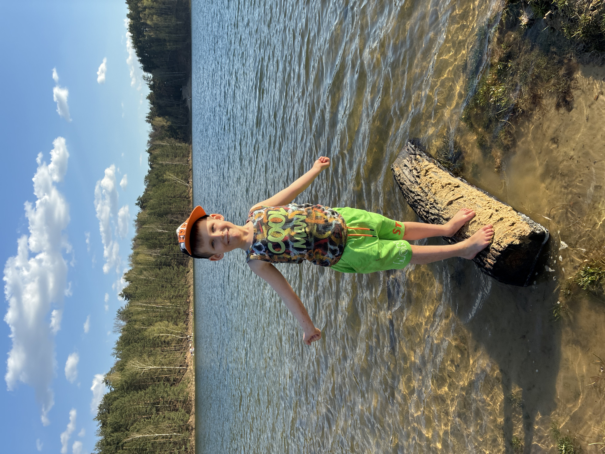 A young boy wearing a colorful tank top, green shorts, and an orange cap stands barefoot on a log in shallow water near a shoreline. The water is clear enough to see the bottom. Behind the boy is a lake and a forest. The sky is blue with scattered clouds. The location is unknown.