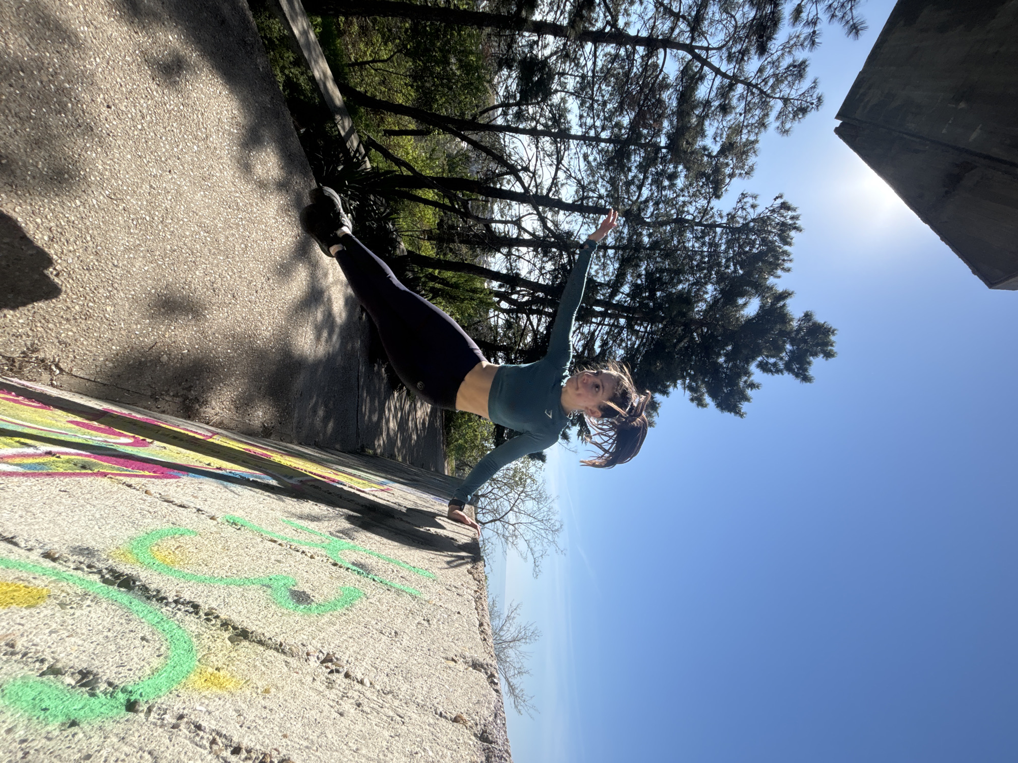 The image depicts a young woman striking a dynamic pose outdoors on a bright, sunny day. She is positioned horizontally against what appears to be a concrete wall or a steep incline, creating an illusion of defying gravity. She wears a dark teal long-sleeved crop top, dark leggings, and black athletic shoes, with her dark hair tied back in a ponytail. Her left hand is planted on the concrete for support, while her right arm is extended outwards and slightly upwards. Her body is held straight and parallel to the surface, and she looks towards the viewer with a focused yet slight smile.
The setting includes a clear blue sky, with the sun casting a bright glare in the upper right quadrant of the image. Silhouettes of trees with both dense and sparse foliage are visible against the sky, suggesting a natural or park-like environment adjacent to a man-made structure. The concrete surface beneath her feet and supporting her hand is textured and features colorful, abstract graffiti in shades of green, yellow, pink, and purple.
The photo is taken from a low-angle perspective, emphasizing the dramatic, gravity-defying nature of the pose and making the ground appear as a vertical wall. The time of day is clearly midday or late morning due to the intense sunlight. The activity suggests an athletic feat, possibly related to parkour, acrobatics, or a creative photoshoot showcasing balance and strength. On her crop top, the brand name "GYMSHARK" is faintly visible.