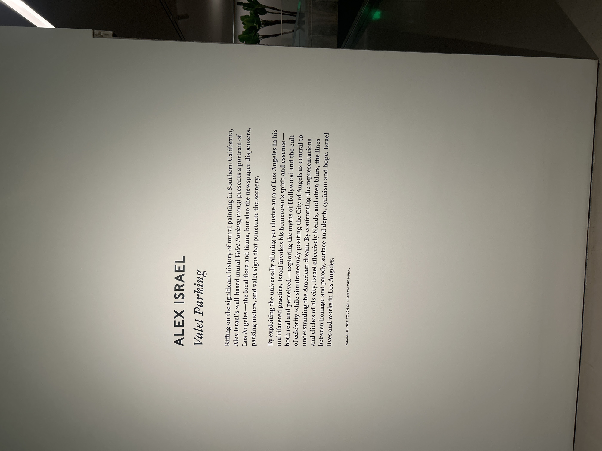 The image shows a sign at an art exhibit for Alex Israel’s “Valet Parking” mural. The text describes the mural as a portrait of Los Angeles. It also mentions the local flora and fauna, newspaper dispensers, parking meters, and valet signs in the scenery. It states that Israel invokes his hometown's spirit and essence while exploring the myths of Hollywood. The sign requests visitors not to touch or lean on the mural. The location is an art gallery or museum in Los Angeles, United States of America.