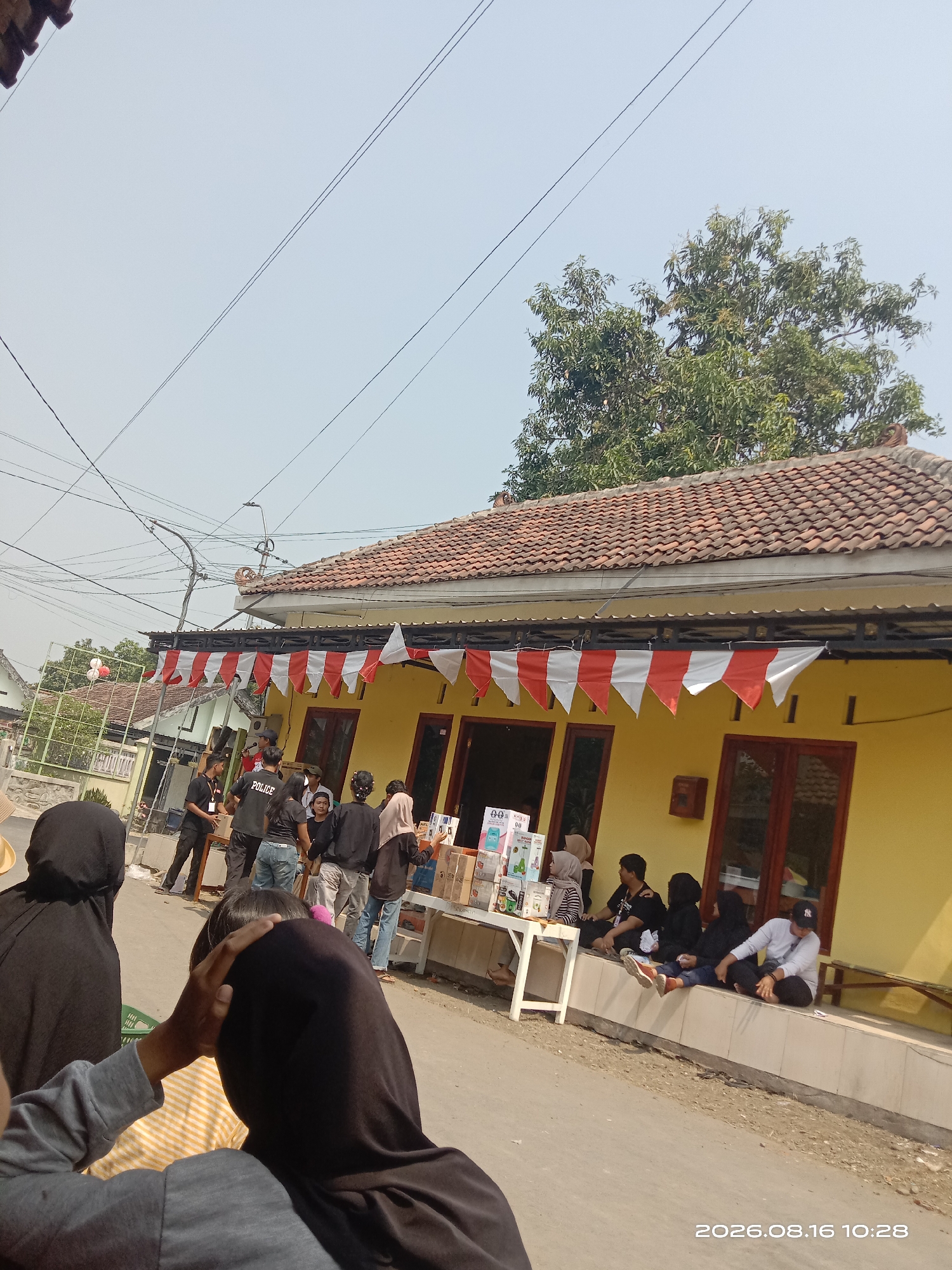 The image depicts a sunny outdoor scene in the city of Banyuarang, Indonesia, likely a residential area given the presence of houses. In the foreground, an individual wearing a black hijab and a grey long-sleeved shirt is partially visible, facing away from the camera and looking towards a group of people.

The central focus is a yellow-painted house with a tiled roof, decorated with red and white pennant flags. In front of the house, a white table displays various boxes, possibly merchandise for sale. Several individuals are gathered around this table and sitting on a low wall in front of the house. Some appear to be interacting with the products on the table, while others are seated, conversing, or observing.

A few individuals are dressed in dark clothing, including some wearing black headscarves. One person in the group is wearing a shirt that says "POLICE". The overall atmosphere appears to be a casual gathering or a small outdoor event. The environment is characterized by a paved road in the foreground, a building with a distinct yellow facade, and trees in the background, suggesting a tropical or subtropical climate. The sky is clear and bright, indicating daylight hours. Electrical wires are visible crisscrossing the upper part of the frame, a common sight in urban and suburban areas.