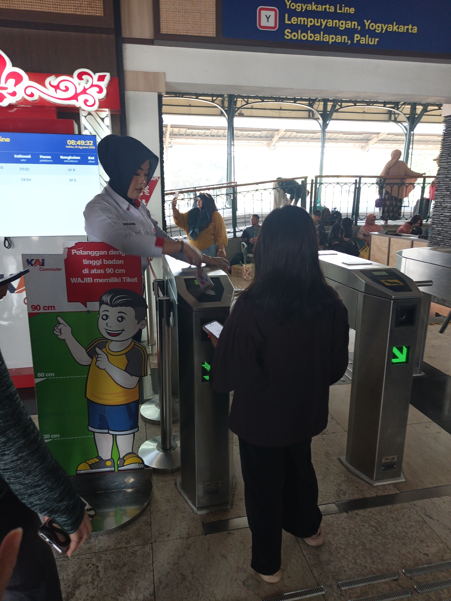 The image depicts the interior of a train station, likely in Klaten, Indonesia, given the signage indicating "Yogyakarta Line Lempuyangan, Yogyakarta, Solobalapan, Palur." In the foreground, a woman in a grey uniform with a black hijab is attending to a turnstile. She is looking down and appears to be assisting a passenger or operating the turnstile. To her right, a person is walking through the turnstile. To her left, a poster is displayed featuring a cartoon character and text related to height requirements for ticket holders. The background shows other passengers waiting and platforms. The environment appears to be a modern, clean, and busy transportation hub.