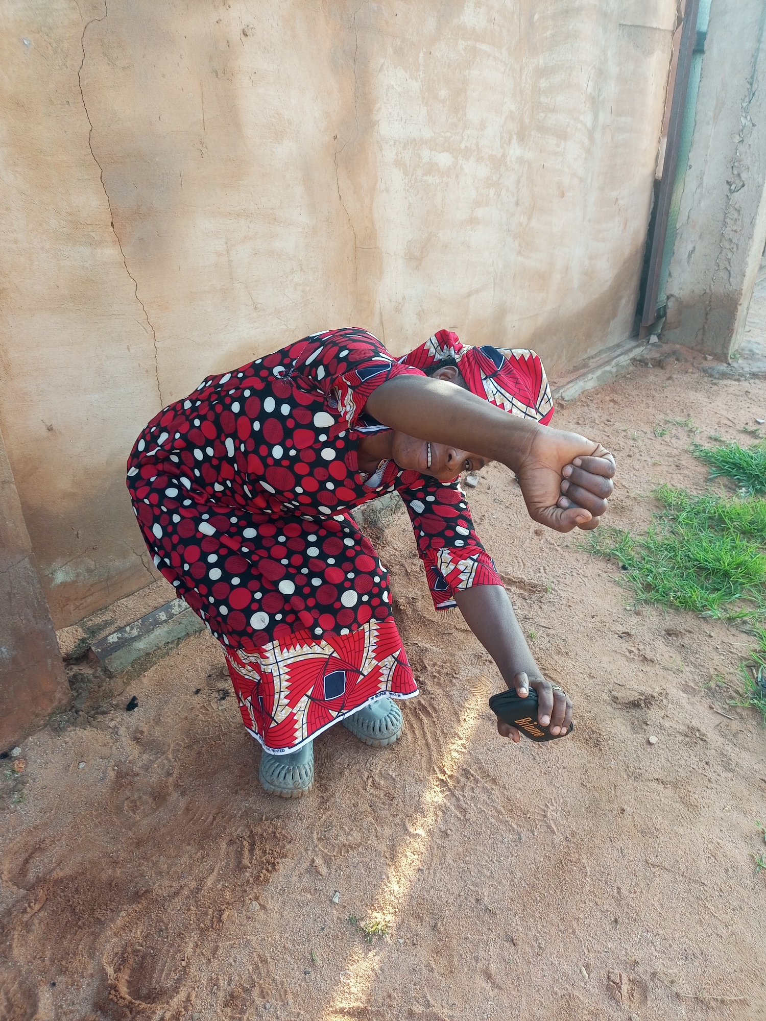 In Tempure, Nigeria, a person wearing a red and black patterned dress and headscarf is bent over, facing the viewer. Their right arm is extended, and their fist is clenched. In their left hand, they are holding a small black object with an orange logo. The person is standing on sandy ground, with a textured, light-colored wall behind them. To the right of the frame, some green grass is visible.