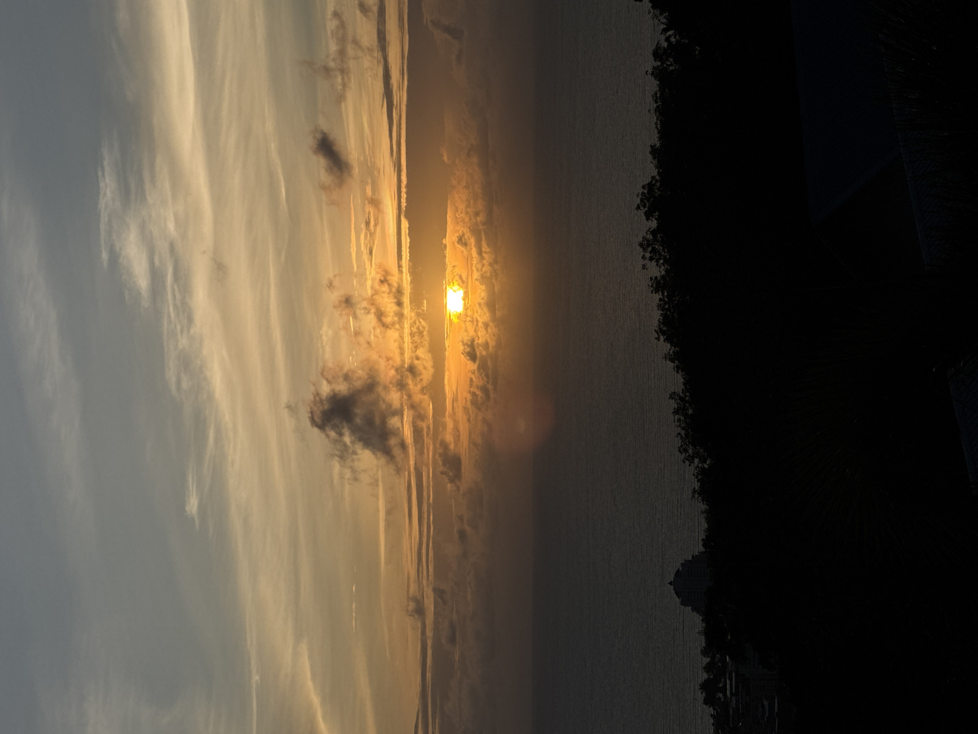 The image depicts a sunset over the ocean, viewed from an elevated position in Patong, Thailand. The sky is filled with hues of orange, yellow, and gray, with wispy clouds scattered across. The sun, a bright orange orb, is partially obscured by clouds near the horizon. The ocean is a deep blue-gray, with gentle ripples. In the foreground, the dark silhouette of lush vegetation and trees lines a ridge, with a distinct, tiered building visible in the mid-ground, partially silhouetted against the fading light. The rest of the city is largely obscured by shadow. There are no people or animals visible in the frame. The scene captures the tranquil beauty of a tropical sunset.