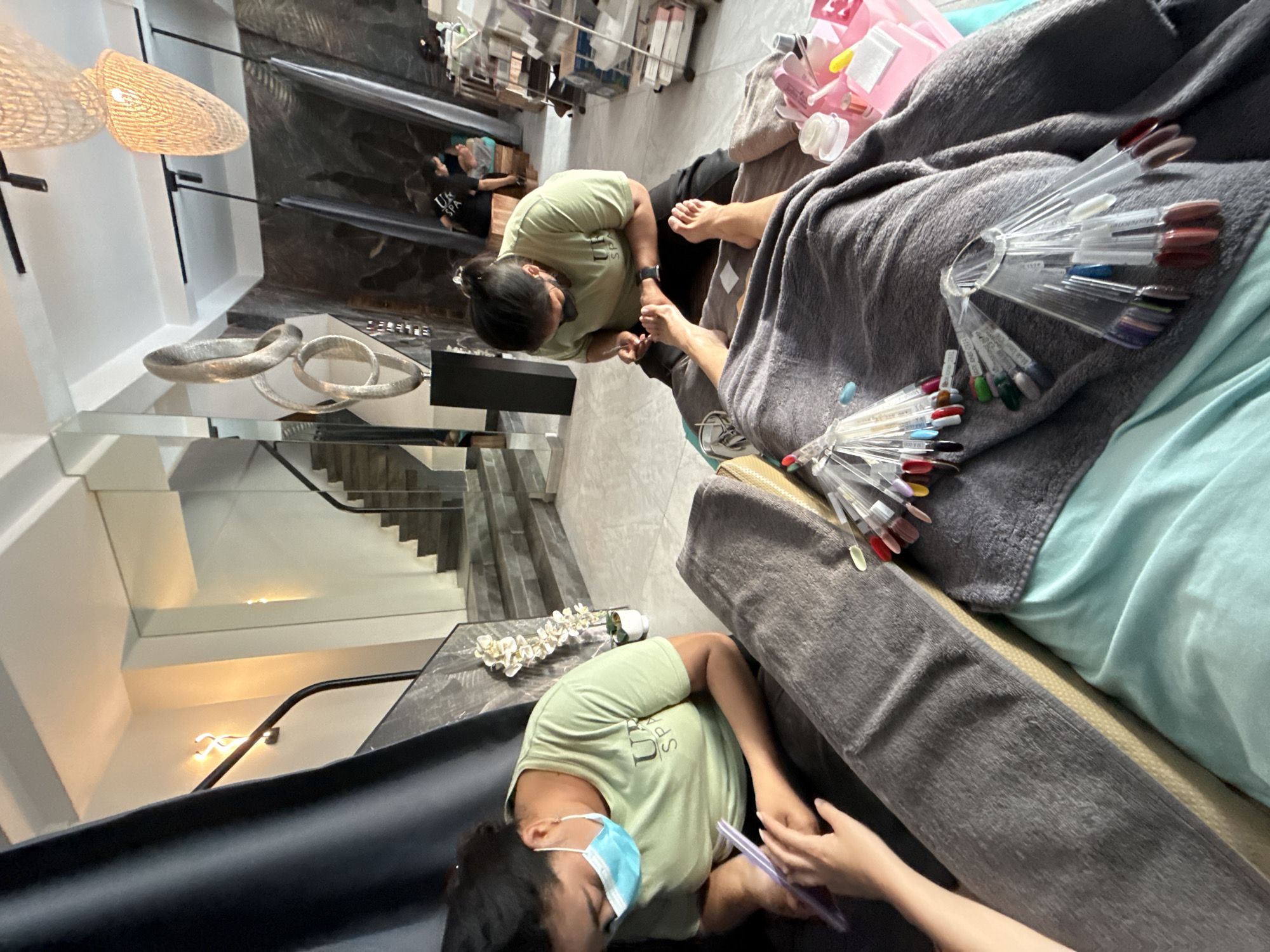 The image depicts a nail salon, likely located in Denpasar, Indonesia, where two individuals are receiving pedicures. One person, wearing a light green t-shirt and a light blue face mask, is having their toenails worked on by a nail technician, also wearing a light green t-shirt and a face mask. The technician is kneeling and appears to be filing or buffing the client's toenails. The client's feet are resting on a gray blanket. Several nail polish swatches are displayed on a stand next to the client. In the background, a set of stairs with a black railing leads up to a higher level. Another person is visible in the far background, also seated. The salon interior features modern decor, including large mirrors and abstract sculptures. The lighting appears to be artificial, with pendant lamps hanging from the ceiling.