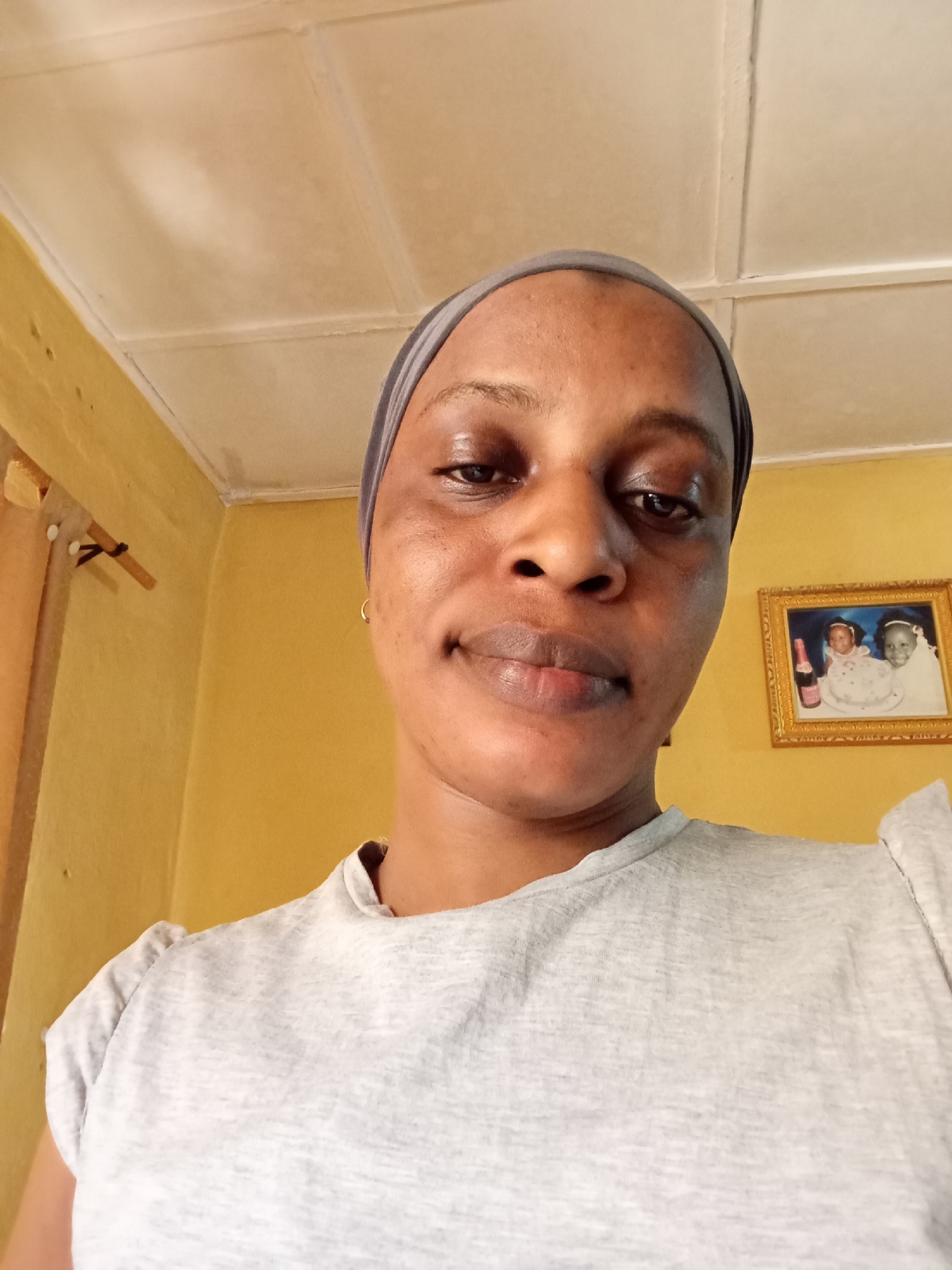 A medium close-up shot depicts a woman indoors in Narayi, Nigeria. She has brown skin, dark eyes, and is wearing a gray headscarf and a gray t-shirt. Behind her, the yellow-painted wall shows a framed picture. Also visible are a wooden window frame and a white ceiling.