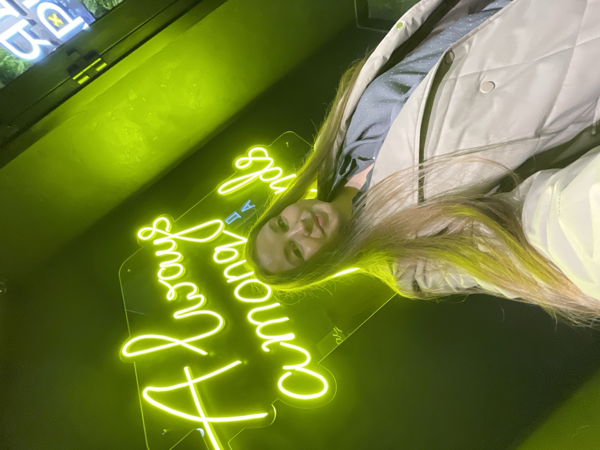 A young woman is standing in front of a neon sign that reads "Always Among," as visible on the sign, at the Always Among restaurant, located in Izhevsk, Russia. The woman is wearing a light-colored quilted jacket over a patterned shirt. The background is dimly lit and appears to be part of the restaurant's interior.