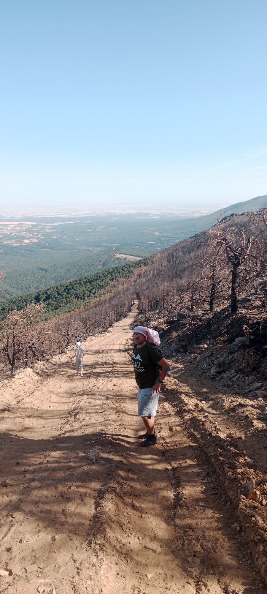 The image captures a scenic view from a mountain trail in Cerezo de Arriba, Spain. A man wearing a green t-shirt, gray shorts, a pink head covering, and glasses is standing on a dirt path. He has a backpack on and is looking upwards. Further down the path, a person dressed in white is walking away from the camera, holding a walking stick. The trail appears to be in a landscape marked by the aftermath of a fire, with numerous blackened, bare trees lining the slopes. Beyond the immediate area, a dense green forest covers the hillside, leading to a vast, hazy landscape with distant plains. The sky is clear and blue.