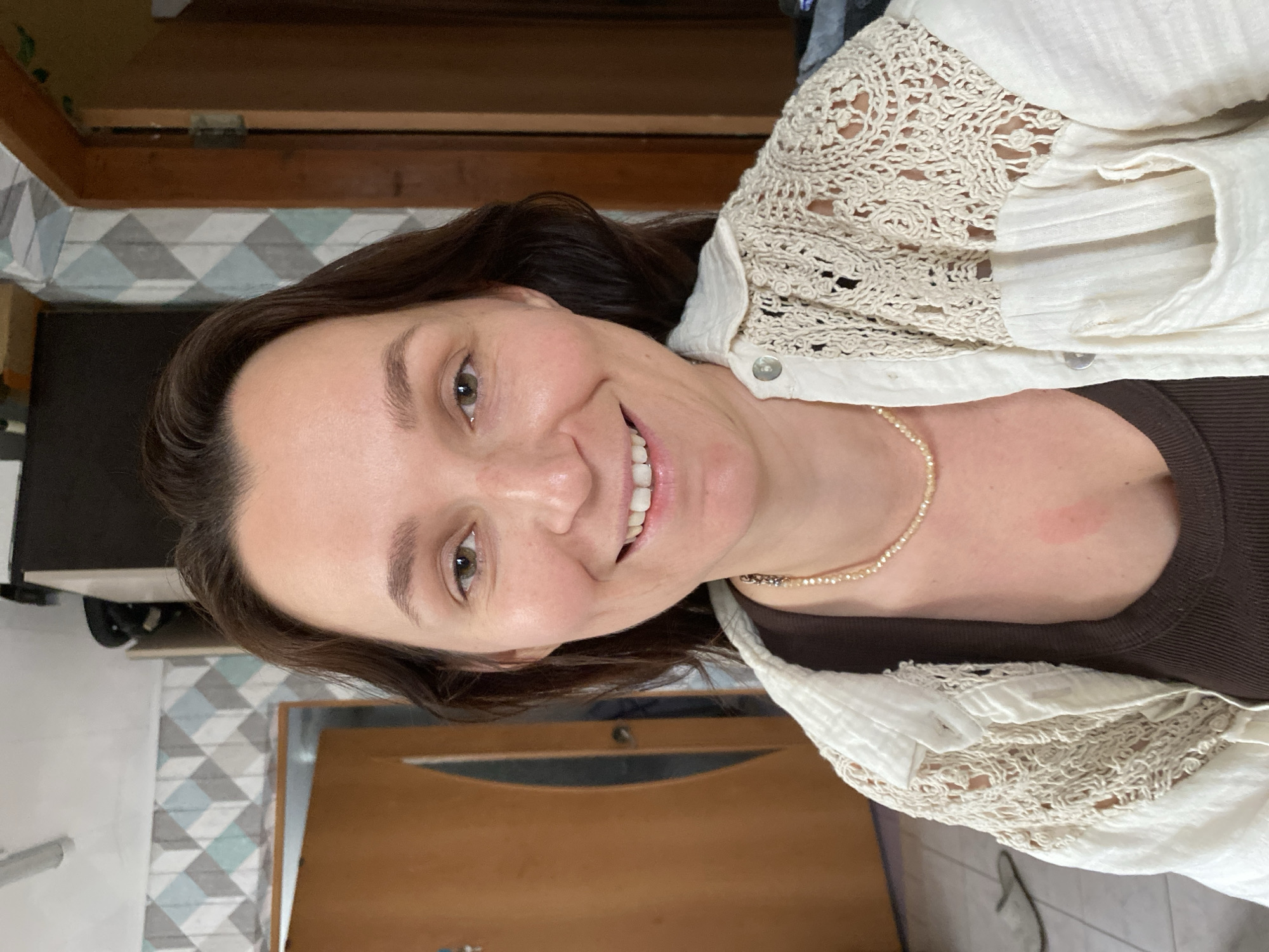 This is a medium close-up, vertically oriented photograph of a woman. She has brown hair and light green eyes, and she is smiling. She is wearing a dark brown top under a cream-colored button-up shirt with lace detailing on the collar and sleeves. A delicate pearl necklace is visible around her neck. There is a reddish mark on her neck, to the right of the necklace.
The background appears to be indoors, with a tiled wall and a wooden door visible. The lighting suggests it is daytime. The overall mood is casual and friendly.
The image has been rotated in processing and needs to be viewed on its side. There is no visible text in the image.
