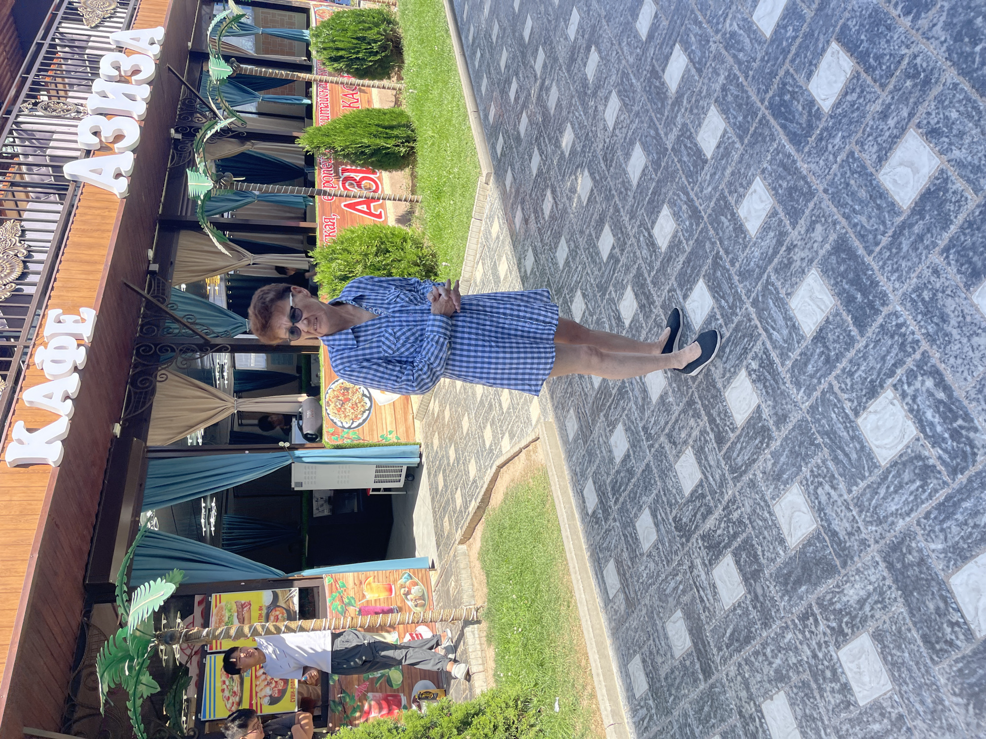 In Bosteri, Kyrgyzstan, a woman with short, styled brown hair and sunglasses stands on a patterned gray and white tiled walkway. She is wearing a blue and white checkered shirt dress and black slip-on shoes. Her hands are clasped in front of her. Behind her, a building with a wooden facade displays large, white Cyrillic lettering that reads "КАФЕ" and "АЗИЗА". The building has an upper level with decorative railings. In front of the building, several artificial palm trees and green shrubs are visible, along with tables and chairs that appear to be set up for outdoor dining. Posters advertising food items like pizza, sushi, and burgers are mounted on the building's exterior. A young man in a white t-shirt and gray pants is standing to the left, near one of the posters. The area appears to be a cafe or restaurant, possibly with a view of a body of water, given the context of Bosteri being a resort town on Issyk-Kul Lake. The sky is a clear blue.