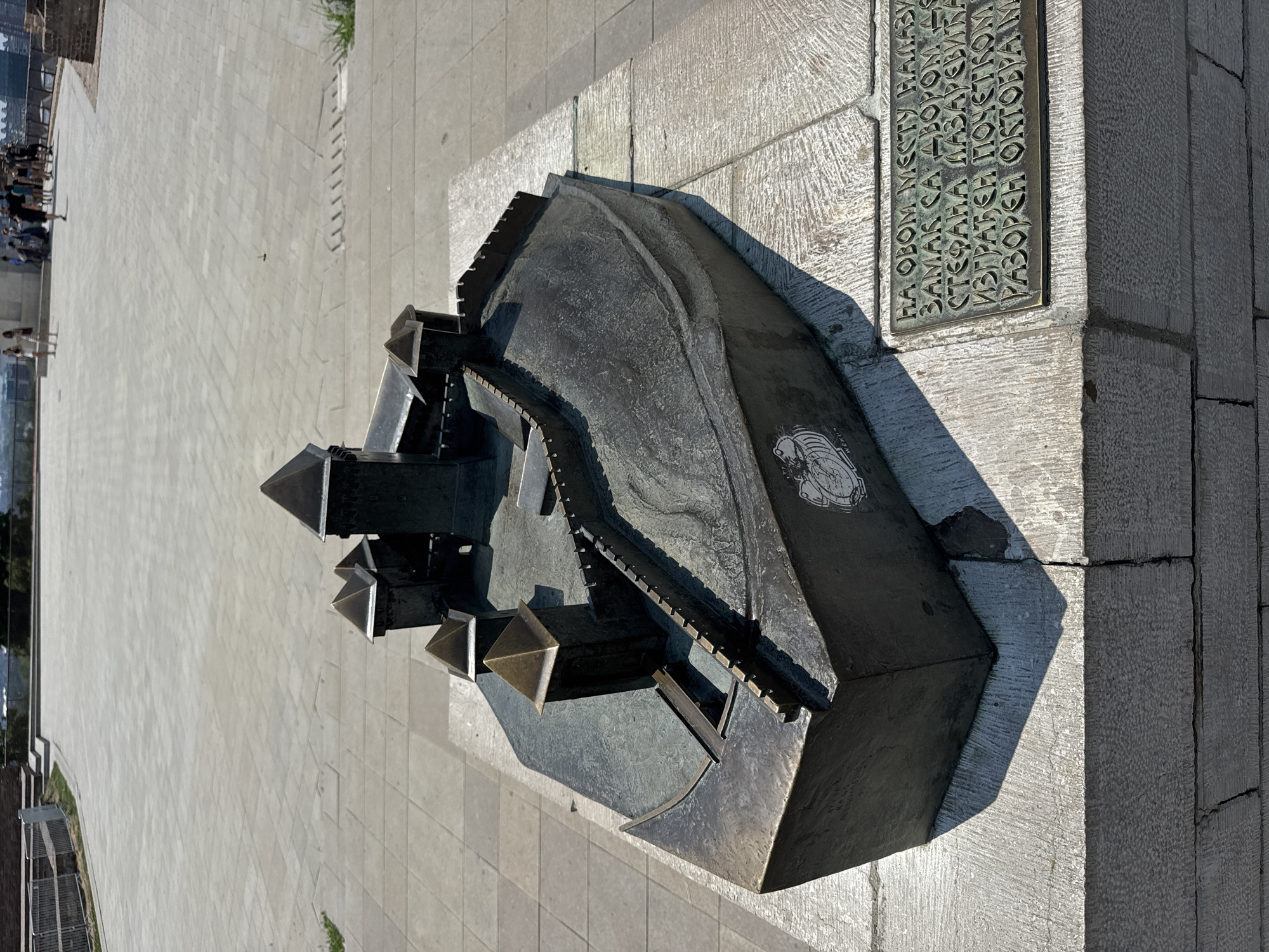 A detailed bronze model of a castle is displayed on a stone pedestal on a paved outdoor area. The model depicts a fortified structure with multiple towers and walls, resting on a sloped surface that suggests a hill or elevation. A plaque with Cyrillic inscriptions is affixed to the front right of the pedestal. In the background, people are visible walking in a larger open area, and beyond them, a cityscape with buildings and a body of water can be seen, indicating an urban setting with a view. The scene appears to be a public space, likely a historical site or viewpoint in Belgrade, Serbia. The lighting suggests it is daytime, with clear shadows cast by the model and the surrounding structures.