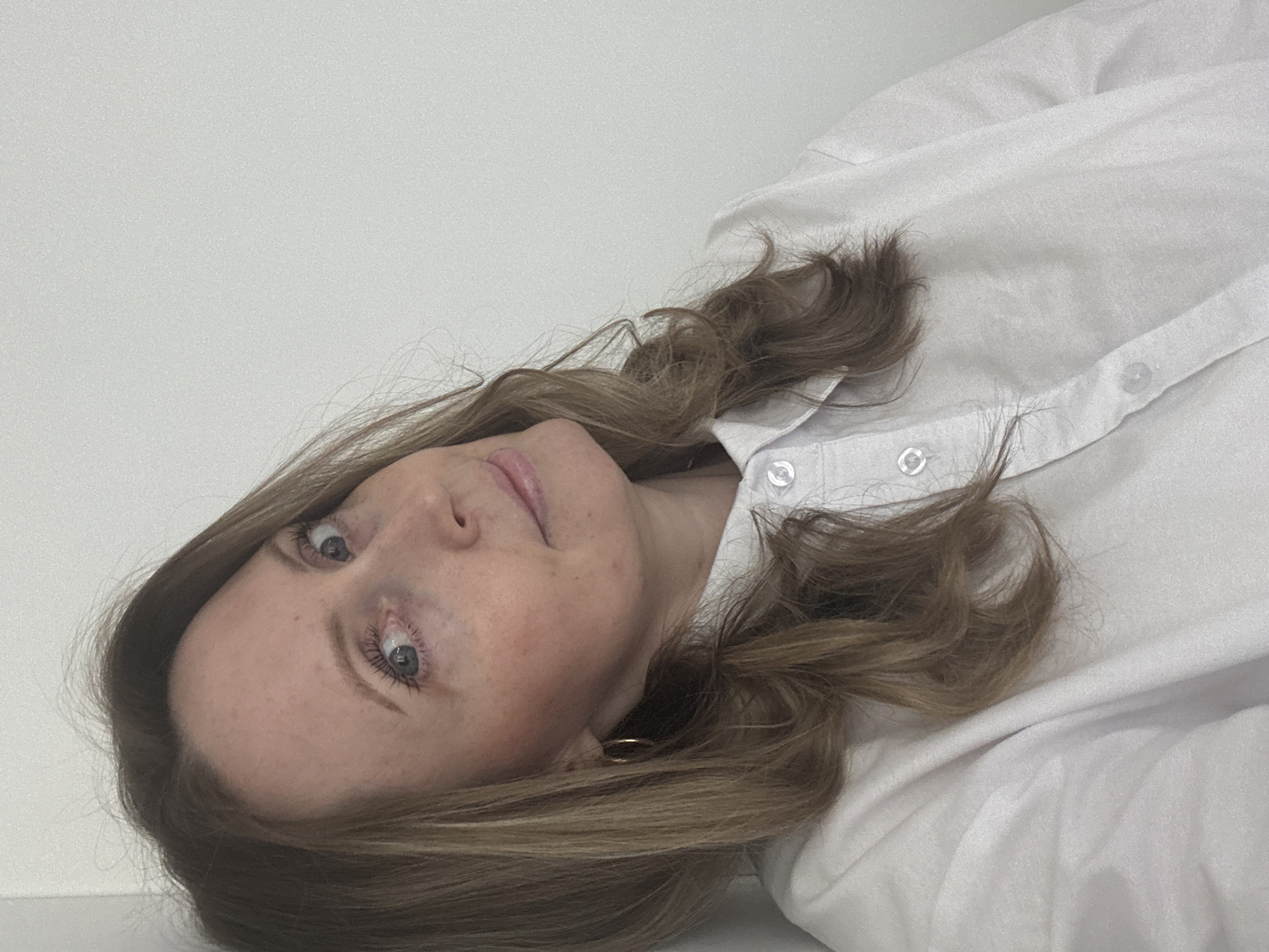 A young woman is featured in a portrait-style photograph, taken in Chaikovsky, Russia. The image is presented with a 90-degree clockwise rotation, showing her head tilted as if she is lying down or leaning against a plain white background. She looks directly towards the camera with a calm, neutral expression.
Her light brown, wavy hair is spread around her head and shoulders. She has fair skin with visible freckles, and light-colored eyes, with a hint of mascara on her eyelashes. She is wearing a white collared shirt with several buttons visible down the front. A small, golden hoop earring is visible on her right ear. The background is an unadorned white wall or surface, creating a minimalist setting that keeps the focus on the subject. The photograph has a slightly grainy texture. No text is visible in the image.
