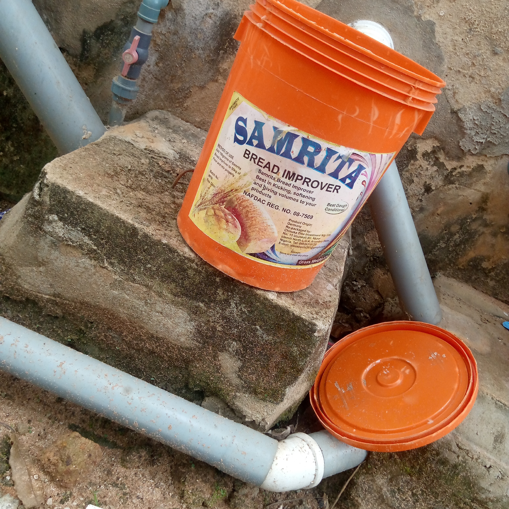 The image presents a still life of objects resting on a concrete structure, likely outdoors. An orange plastic bucket labeled "Samrita Bread Improver" is the focal point. The label indicates the product is manufactured in Germany and re-packaged in Nigeria by Chinaka Dike Investment Nig. Ltd., located in Nkpor, Anambra State. A matching orange lid lies nearby. Gray PVC pipes run through the scene, and one has a valve. The concrete block upon which the objects rest is weathered and stained, suggesting an outdoor environment. Based on the label information, it is likely that this image was taken in or around Nkpor in Anambra State, near the city of Ogidi, Nigeria. The overall impression is one of a utilitarian scene.