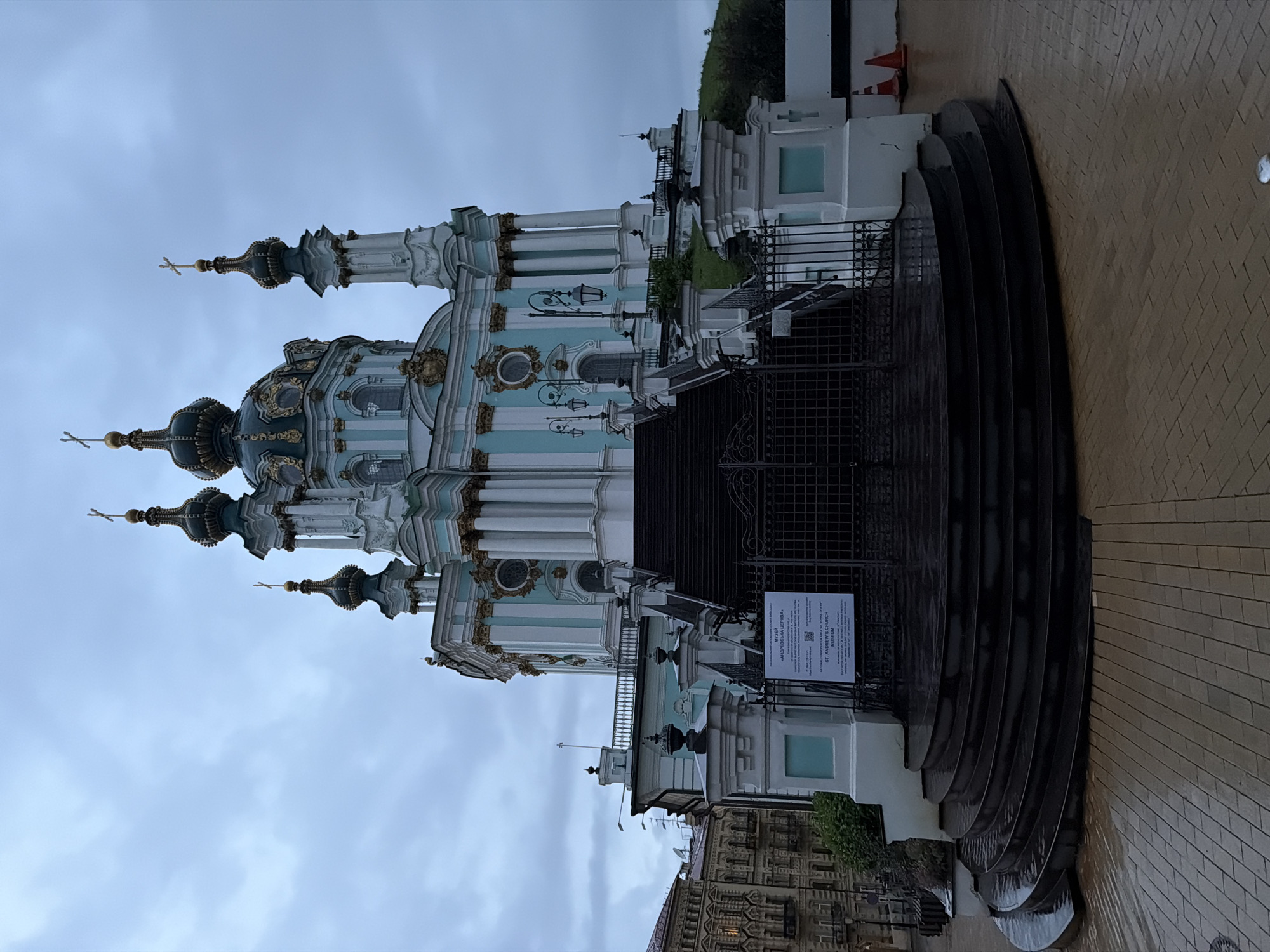 This is a wide-angle, eye-level photograph of Saint Andrew's Church in Kyiv, Ukraine, on a cloudy day. The ornate Baroque church, painted in pastel blue and white with gold accents, dominates the upper portion of the frame. It features multiple domes topped with crosses, classical columns, and decorative elements. A grand, dark, curved staircase with a wrought-iron gate leads up to the church entrance. The steps and surrounding paving stones appear wet, suggesting recent rain. To the left of the church, older, less ornate buildings are visible. In the foreground, the wet, patterned pavement is illuminated by the overcast sky. Two orange traffic cones are placed on the right side of the frame. There are no people or animals visible in the scene. The overall atmosphere is somber due to the cloudy weather.