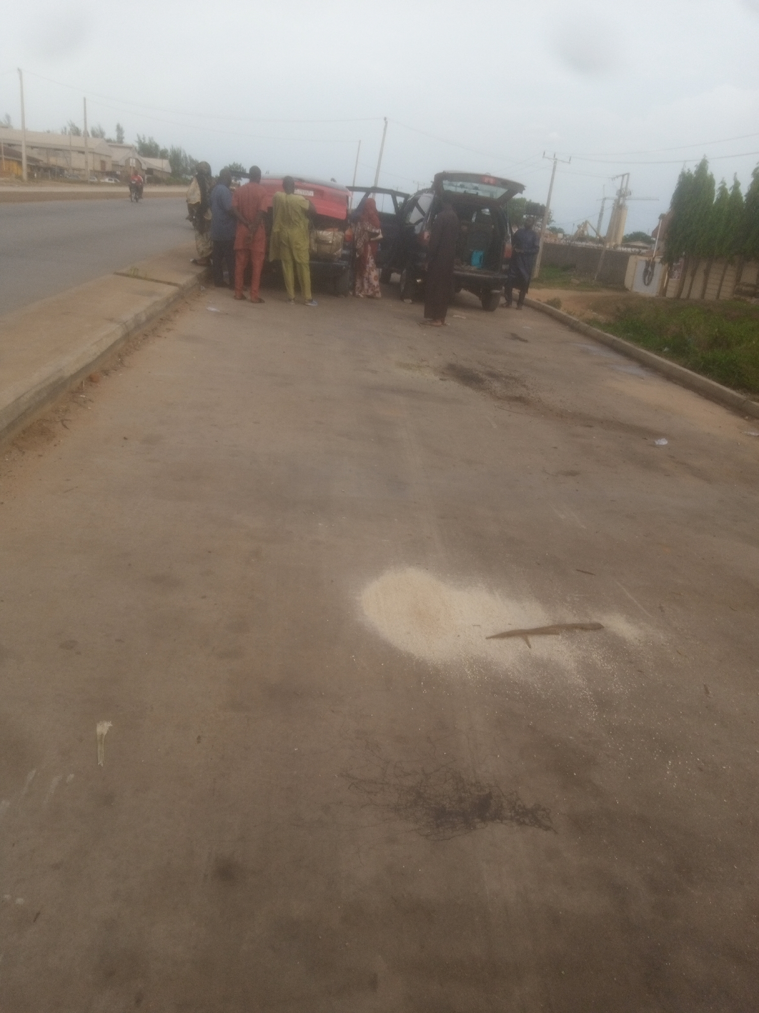 A group of approximately seven individuals are gathered around a red utility vehicle with its rear hatch open. The vehicle is parked on the side of a paved road. Some individuals are standing near the open hatch, and others are standing on the side of the road. The environment appears to be an urban or semi-urban setting, with a road, sidewalk, and some vegetation visible. The sky is overcast. A spill of light-colored granular material, possibly rice, is visible on the paved surface in the foreground, along with some dark debris.
