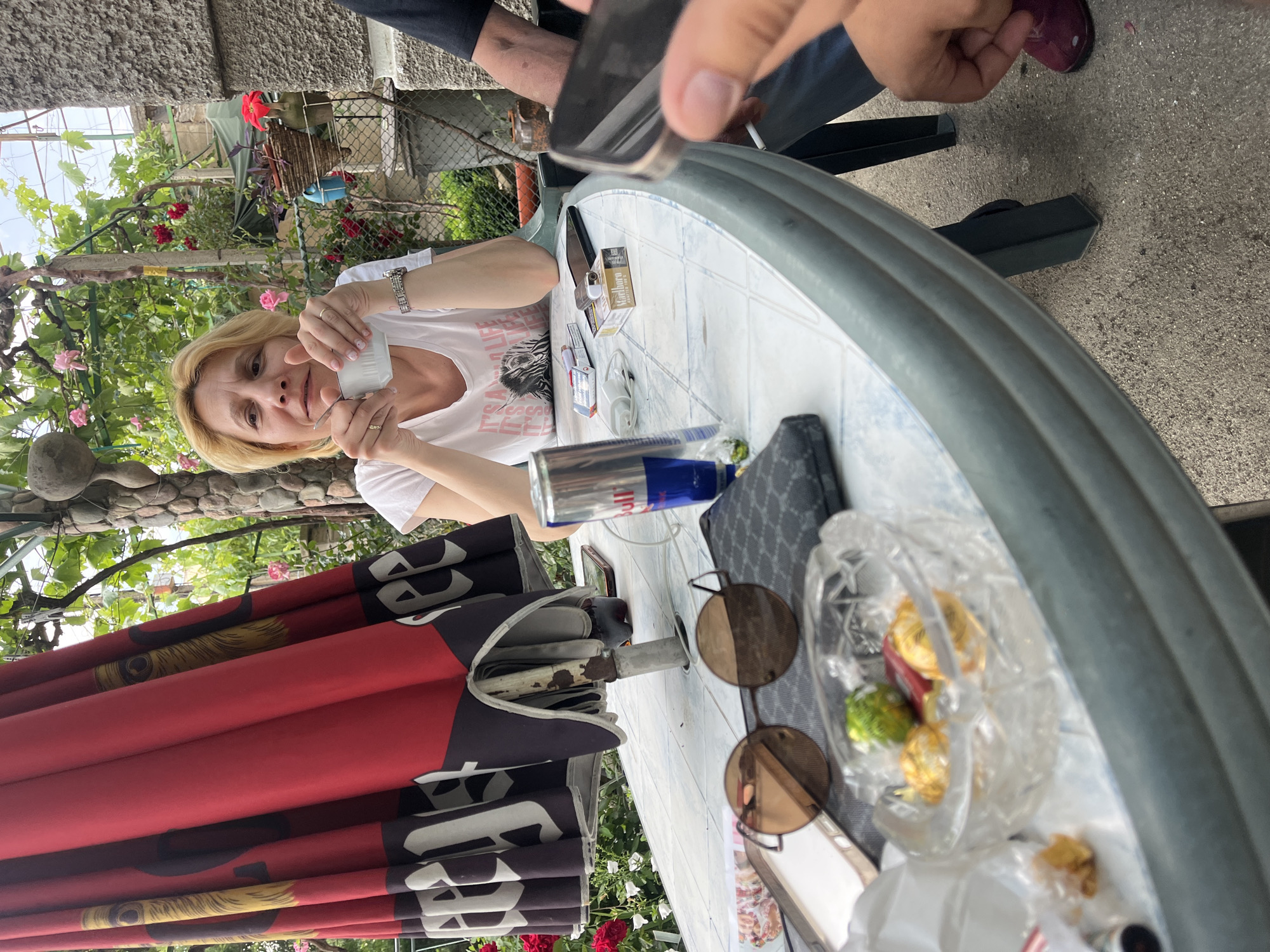 A candid, eye-level photo captures a casual outdoor gathering on a bright day. The main subject is a blonde woman with light skin, wearing a white t-shirt with a zebra graphic and the text "IT'S A LIFE". She holds a white cup with both hands, looking directly at the camera with a neutral expression. She wears a watch on her left wrist and rings on her fingers.
The woman is seated at a white, round plastic table, which is laden with various items. These include a pack of "Marlboro" cigarettes, a lighter, another smaller cigarette pack, a blue and silver "Red Bull" can, a dark-colored smartphone, and what appears to be a charging cable or earbuds. Also on the table are a pair of round, brown-tinted sunglasses, a dark patterned bag or wallet, and a clear glass bowl filled with gold-wrapped chocolates or candies.
In the immediate foreground, another person's hand is visible, holding a black smartphone over the table, suggesting they might be taking a photo or video. A foot in a purple shoe is visible under the table.
The setting appears to be a garden or patio, characterized by lush green foliage, a vine-covered pergola overhead, and blooming pink and red roses. A fence is visible in the background, along with concrete ground and a stone wall structure. To the left of the table, a large, folded red and black umbrella stands upright. The umbrella features stylized faces or figures as part of its design, though specific text or branding is hard to discern due to it being folded. The overall atmosphere suggests a relaxed, social outdoor occasion.