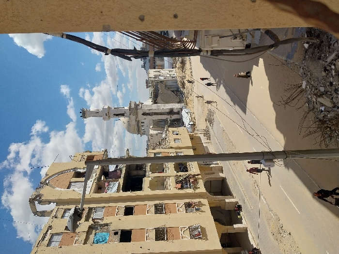 The image depicts a street scene in Khan Yunis, Palestinian Territory, under a partially cloudy blue sky. In the center-left, a severely damaged multi-story apartment building, constructed from light-colored concrete, shows extensive structural compromise with collapsed sections, exposed interiors, and broken windows. Laundry hangs from balconies and damaged sections. A tall grey utility pole with a streetlamp stands in front of this building. In the background, a large mosque with a prominent dome and a tall, multi-tiered minaret is visible, also appearing structurally damaged, with rubble surrounding its base.
The paved street in the foreground and mid-ground is relatively clear but flanked by debris and rubble. Approximately ten individuals are visible walking along the street. In the bottom left foreground, a person pushes a cart. Further up the street, two men walk together, and several other figures are scattered in the mid-distance. On the right side, an external wall section of another building frames the view, with exposed electrical conduits and what appears to be a mounted electrical box or HVAC unit. Piles of concrete debris and twisted rebar are visible in the immediate foreground, indicating widespread destruction.