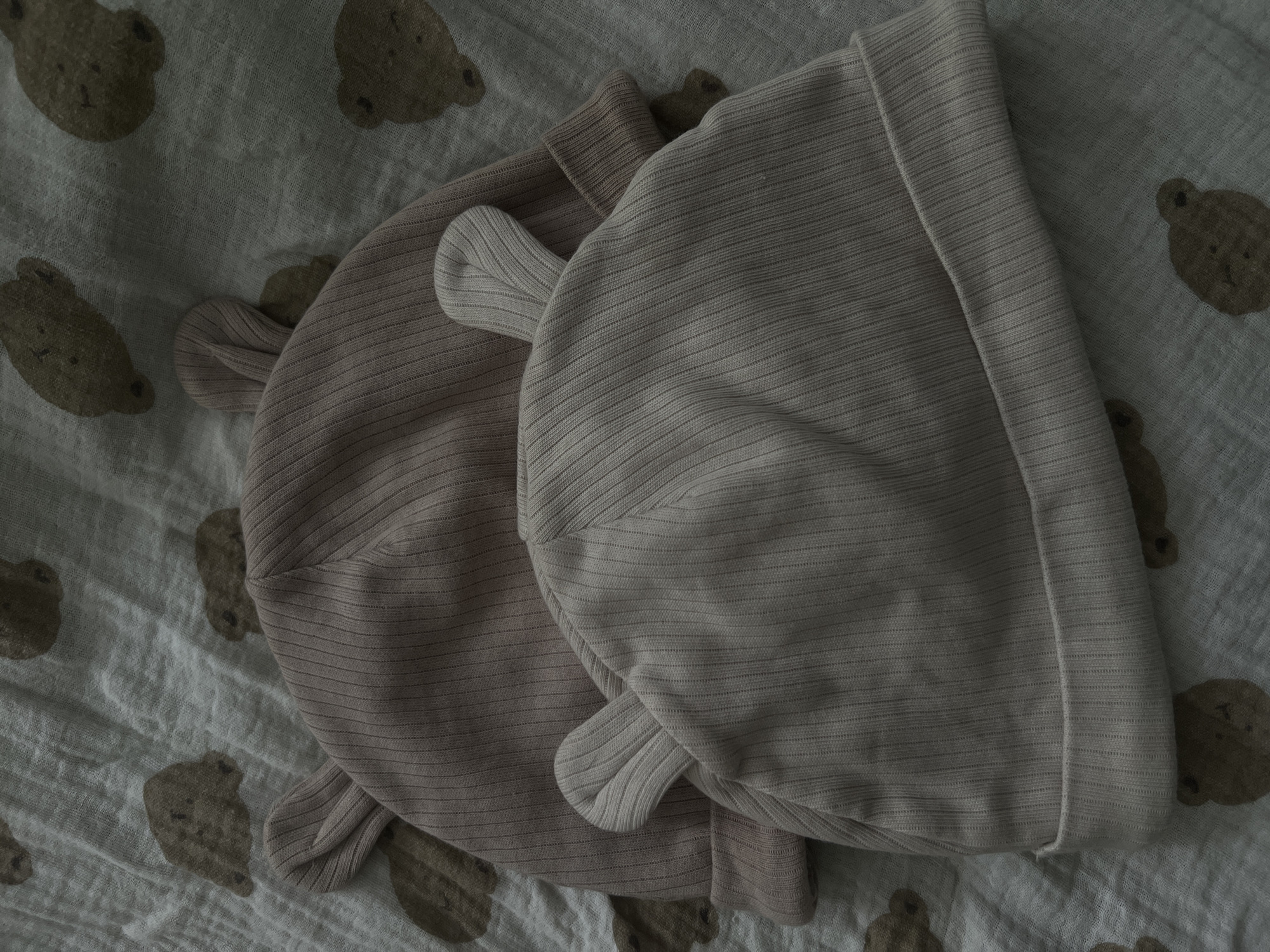 Two ribbed, tan baby hats with attached bear ears are placed on top of each other on a light blue muslin swaddle blanket with a pattern of brown teddy bears. The hats are laid diagonally across the frame, with the bottom hat closer to the viewer. The swaddle blanket is slightly wrinkled and forms the background of the image. The lighting is dim, casting soft shadows.