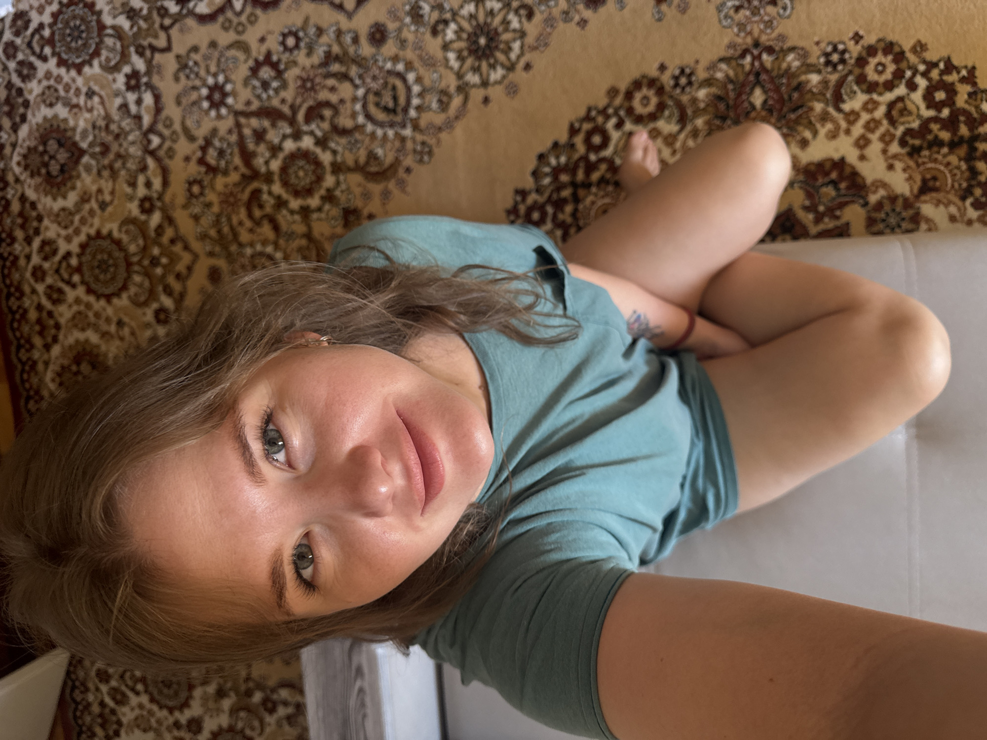 A woman is seated on a white couch in Chaikovsky, Russia. The woman has light brown hair and blue eyes. She is wearing a teal t-shirt. She is seated with her legs crossed. A rug with an elaborate brown, beige, and black pattern is visible in the background.